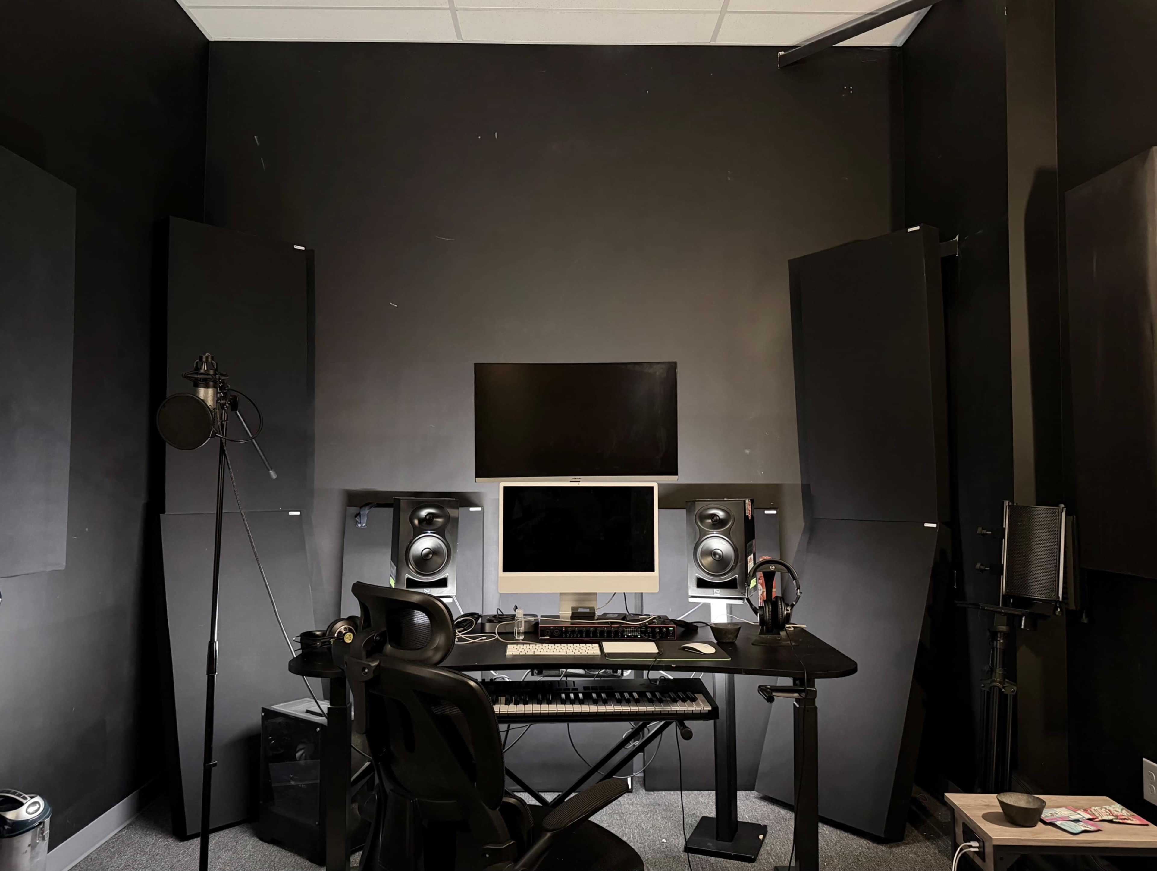 The image shows a music production room featuring a black desk with a computer, studio monitors, a microphone, and soundproofing panels on the walls.