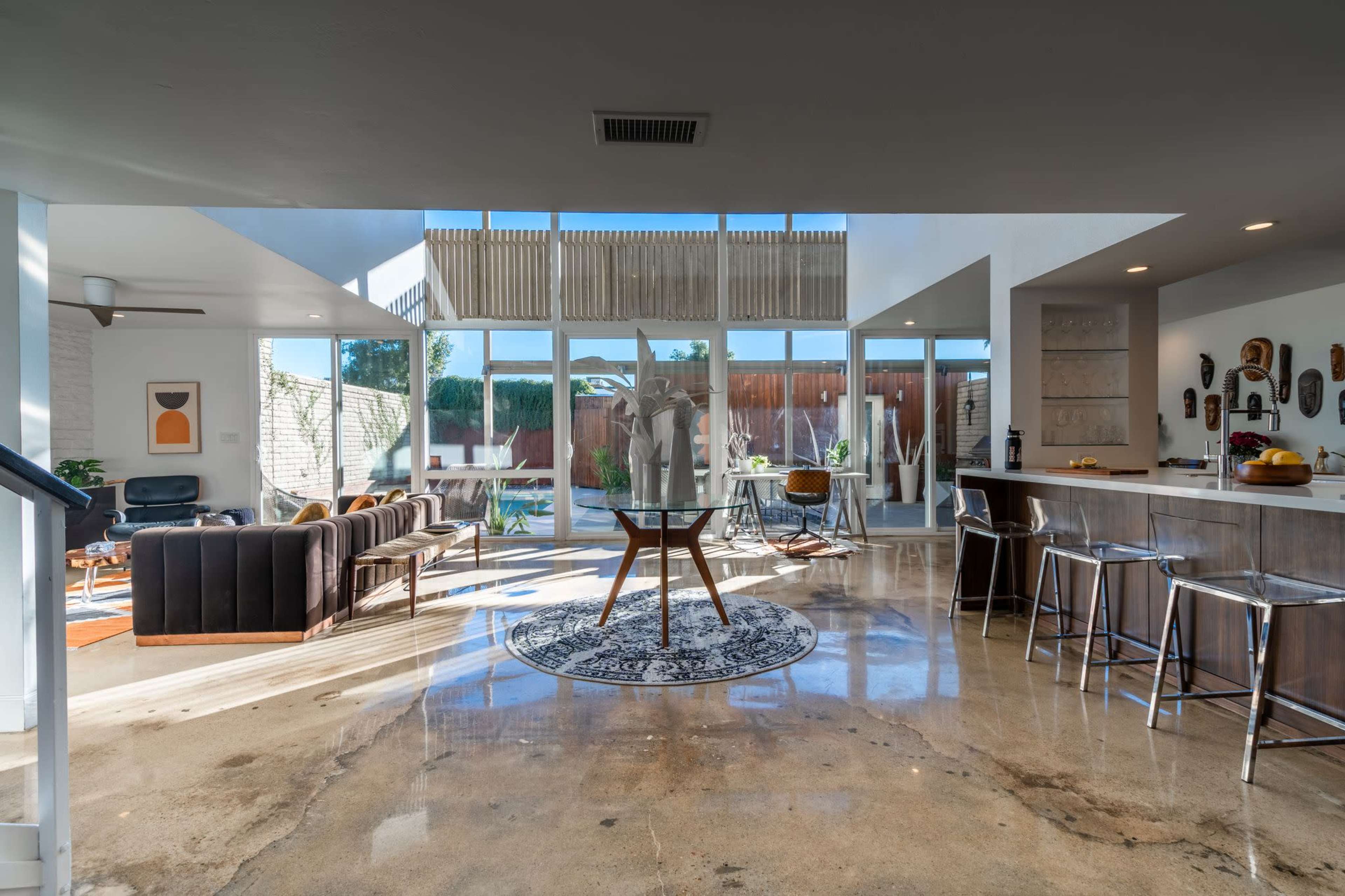 A spacious living area features a modern design with large windows, a dining table, and a kitchen bar, complemented by polished concrete floors.