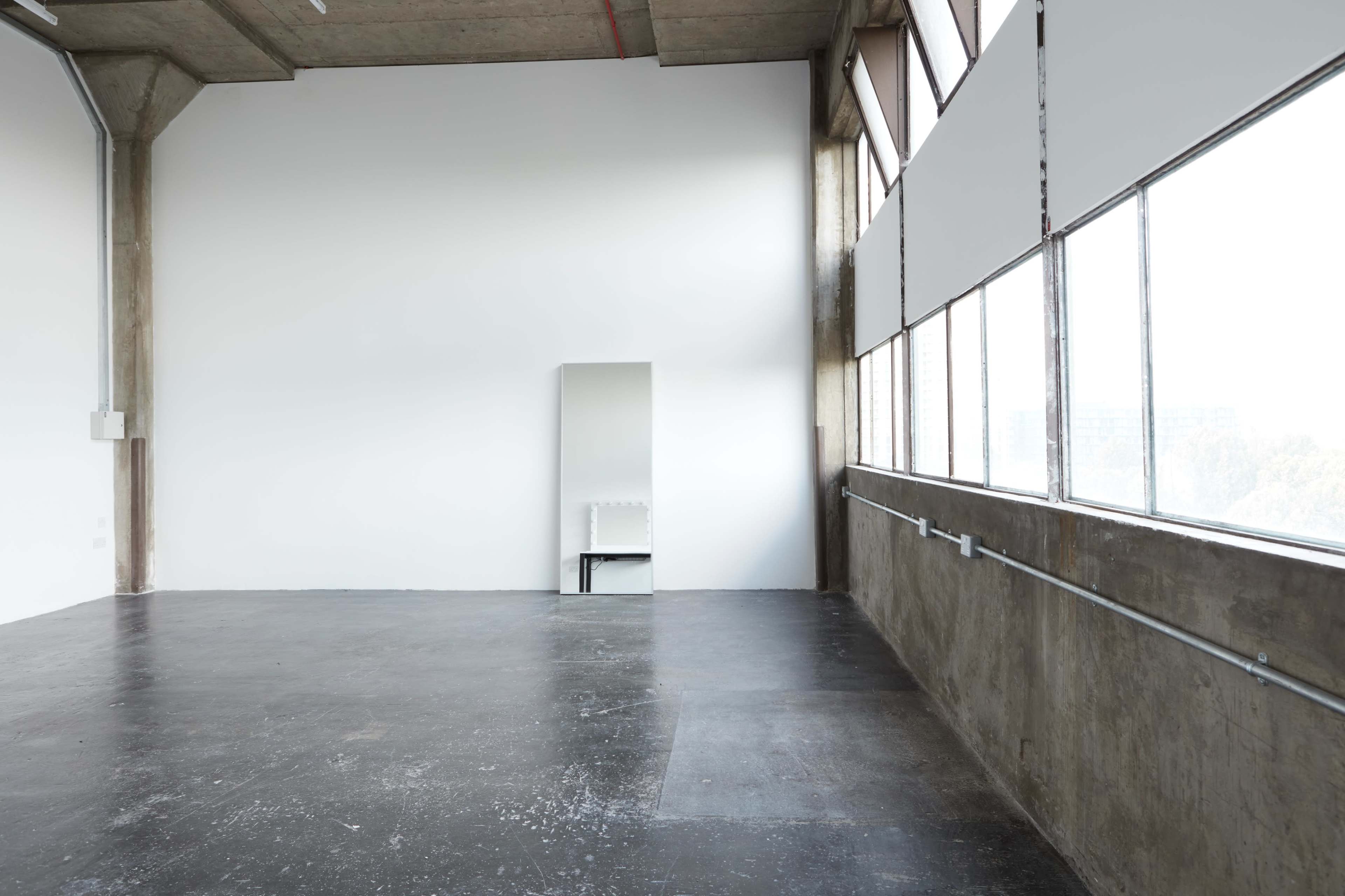 A spacious, industrial-style room with large windows, a concrete ceiling, and a mirrored partition reflecting a chair.
