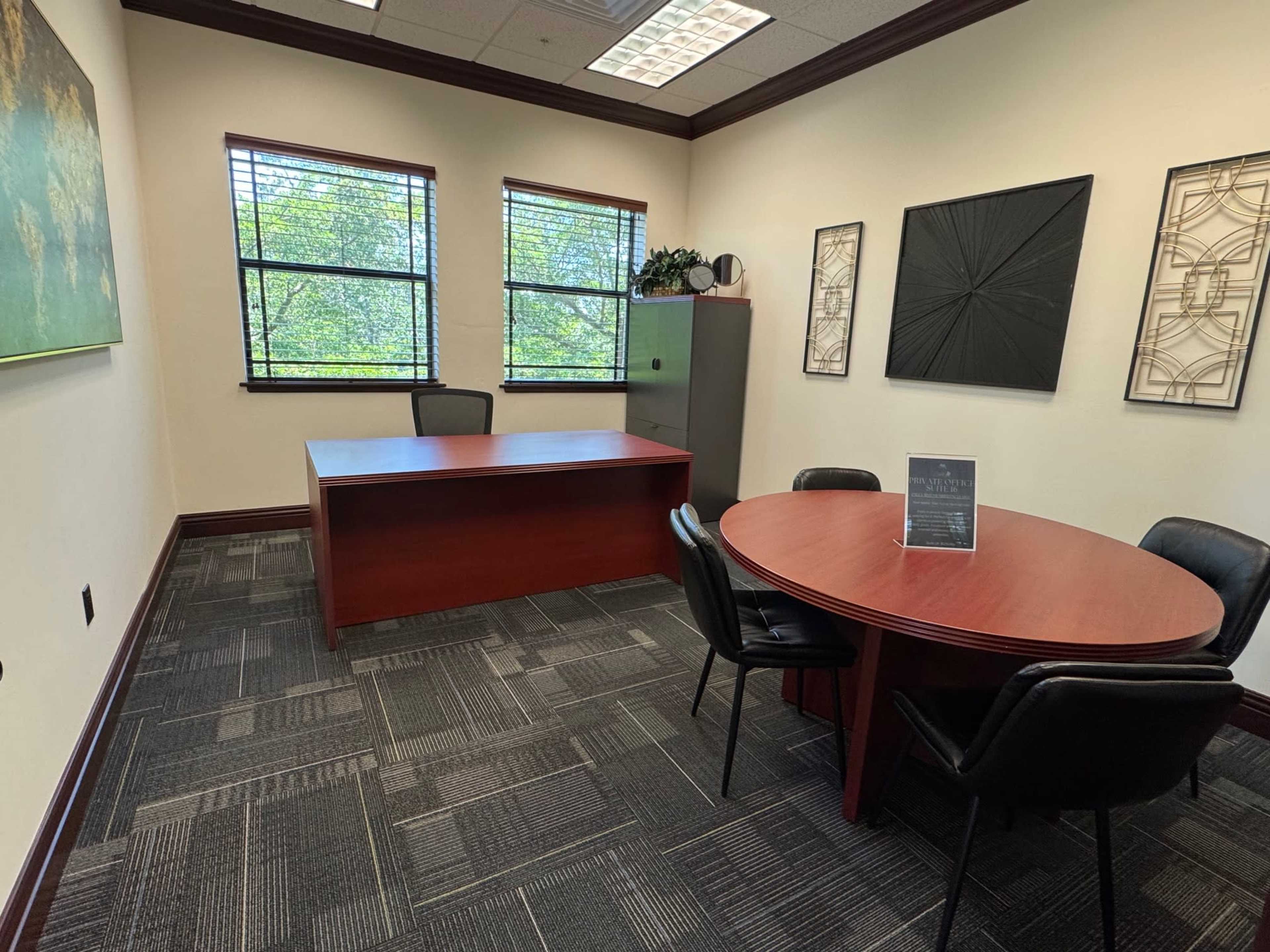 The image shows a modern office space featuring a wooden desk, a round table with chairs, and large windows that provide natural light.