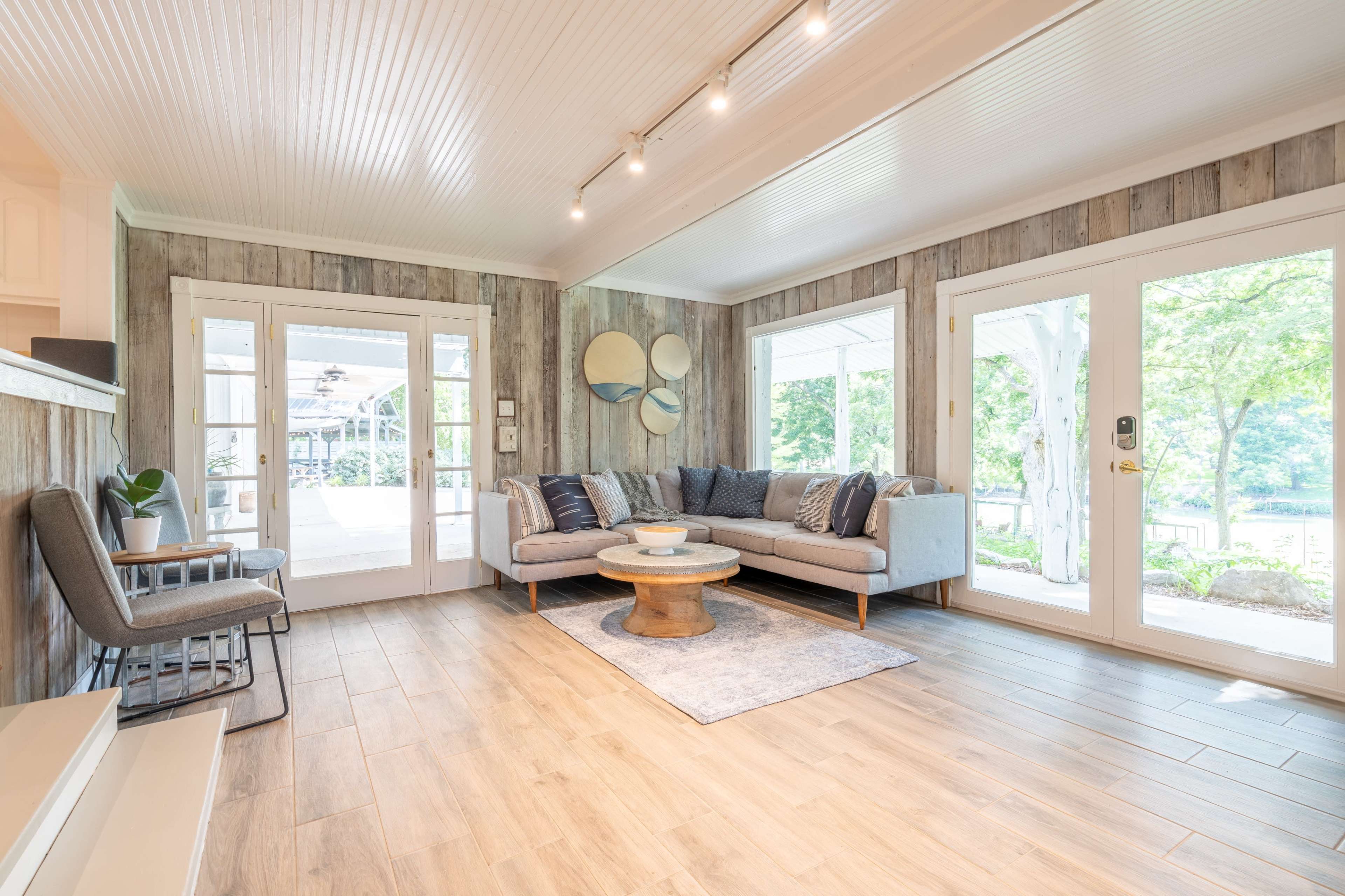 The image shows a bright living room with a gray sectional sofa, a round wooden coffee table, and large glass doors opening to an outdoor patio area.