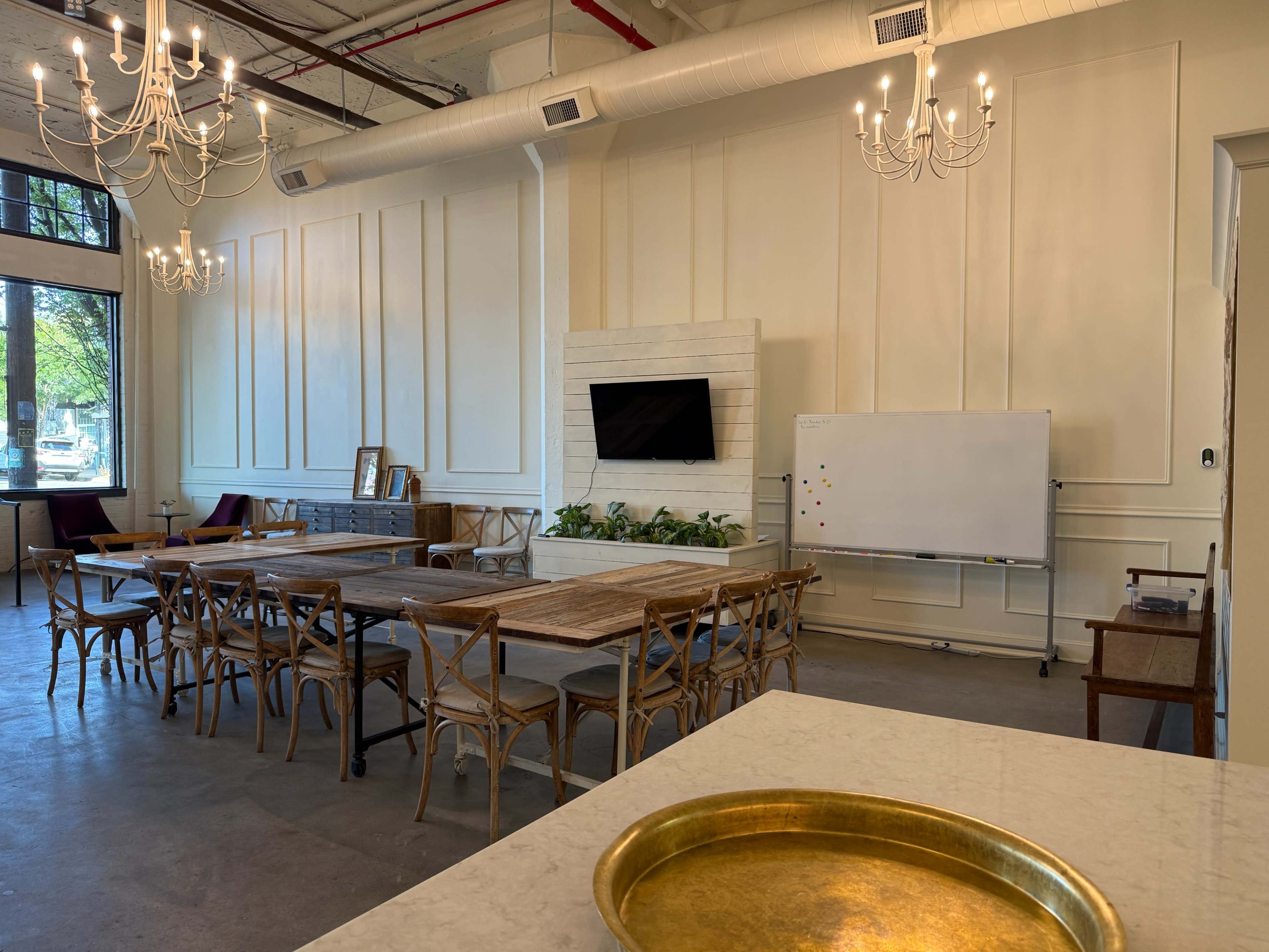 The image shows a spacious room with wooden tables and chairs, a wall-mounted TV, a whiteboard, and decorative chandeliers.