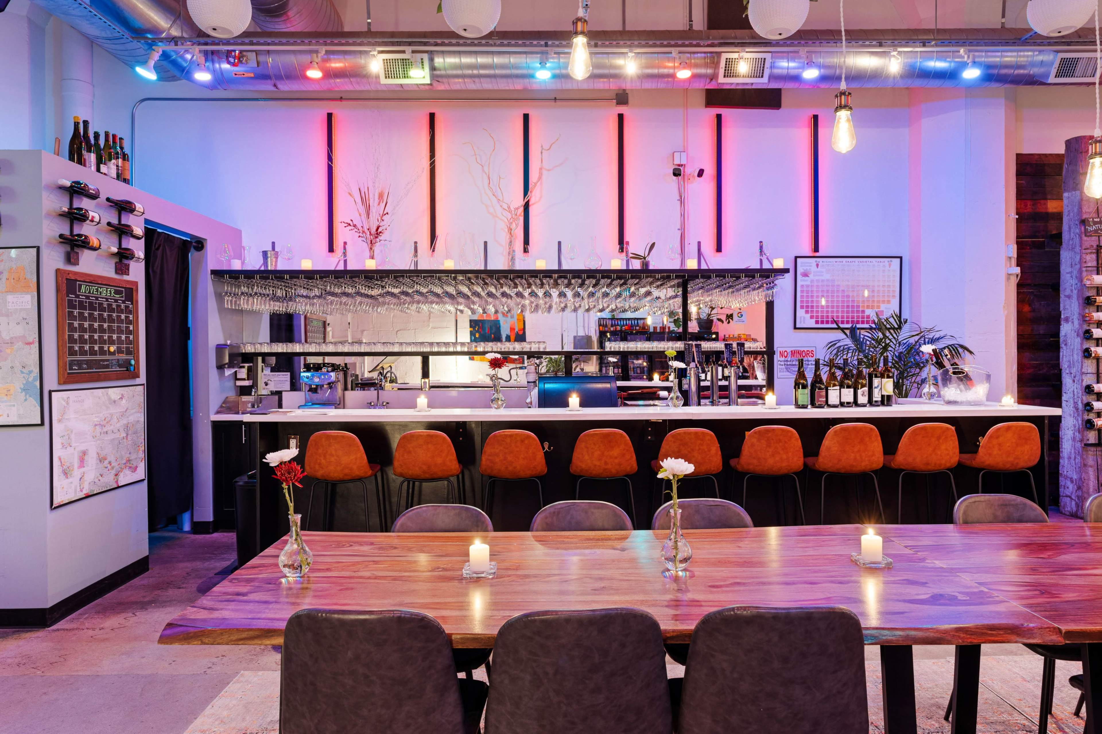 A modern bar area with a long table set for dining, illuminated by colorful lights and decorated with flowers and candles.