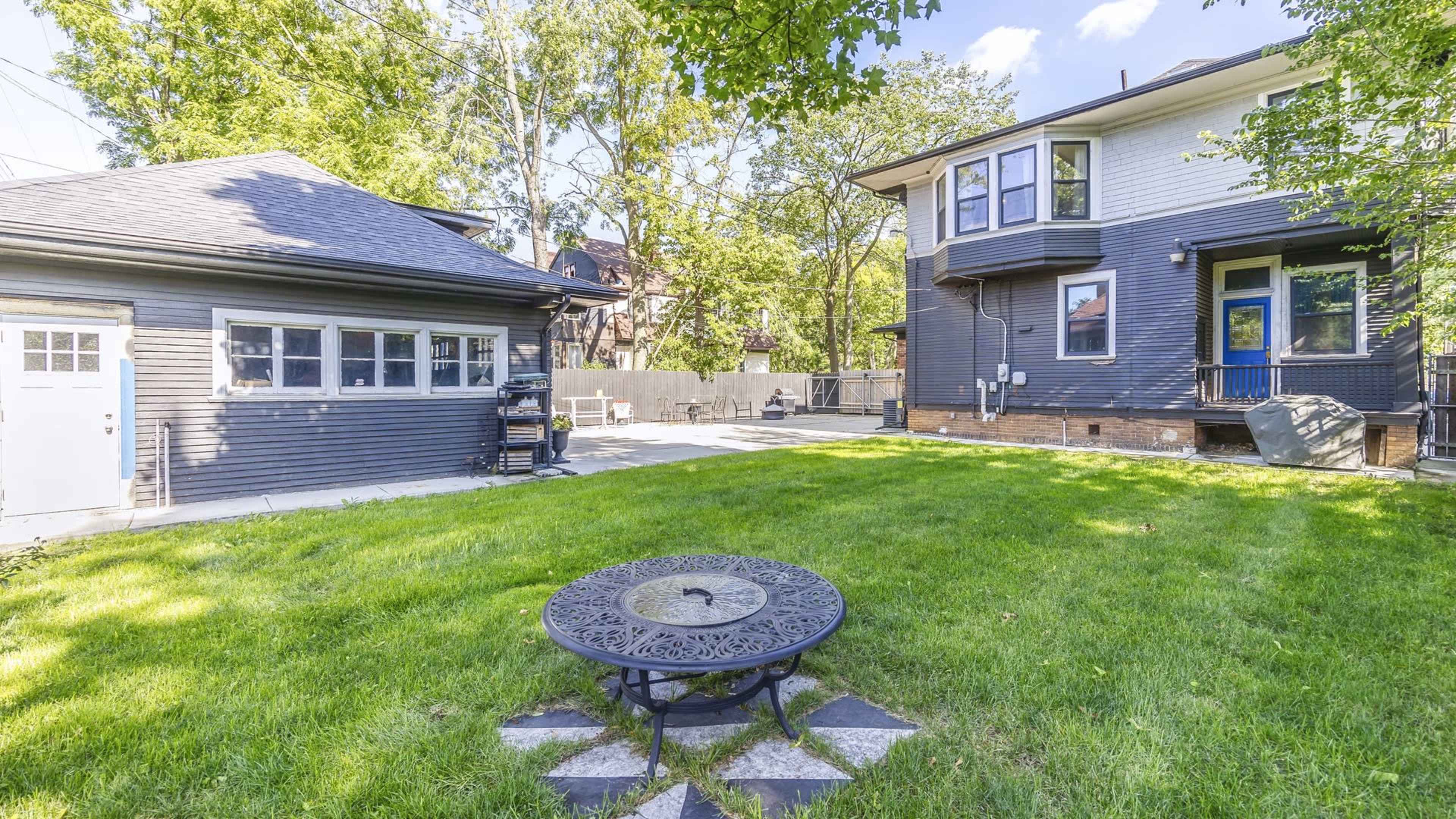A backyard featuring a circular metal table on a stone patio, surrounded by a green lawn and a two-story house with a deck and a separate garage.