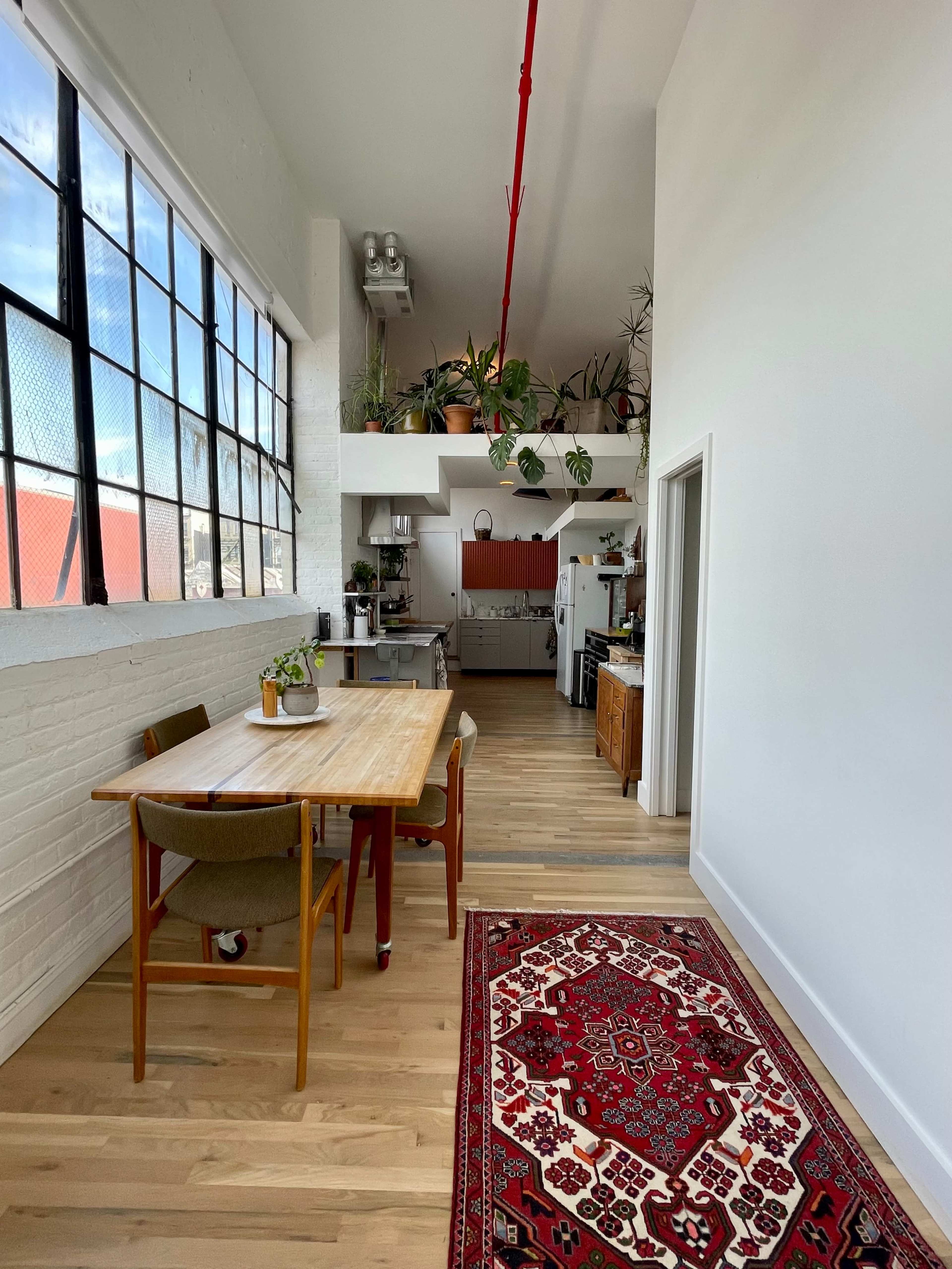 The image features a well-lit, modern interior with a dining table and chairs near a large window, facing an open kitchen adorned with plants and wooden elements.