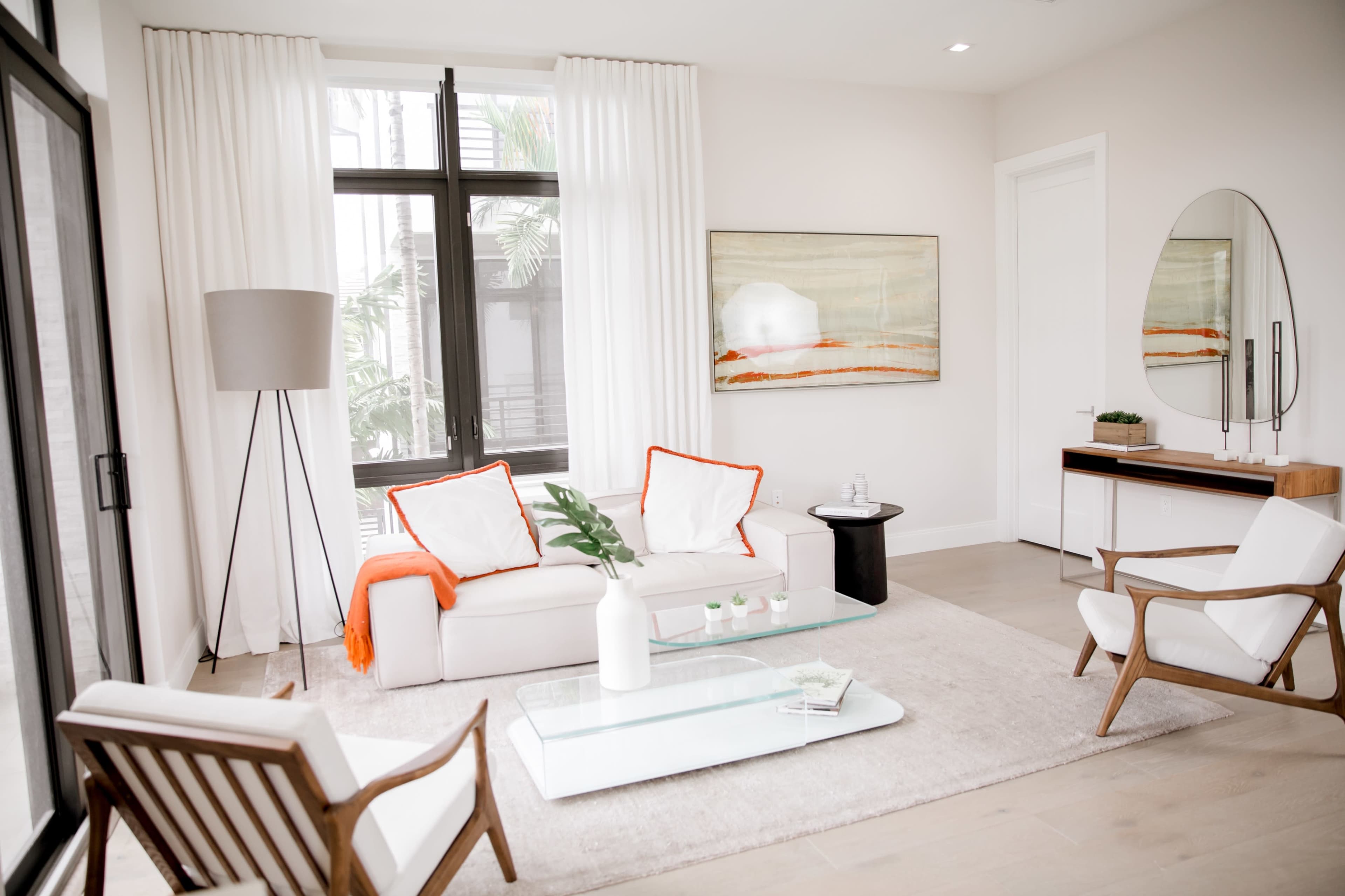 A modern living room with a white sectional sofa, two white chairs, a glass coffee table, and a large abstract painting on the wall.