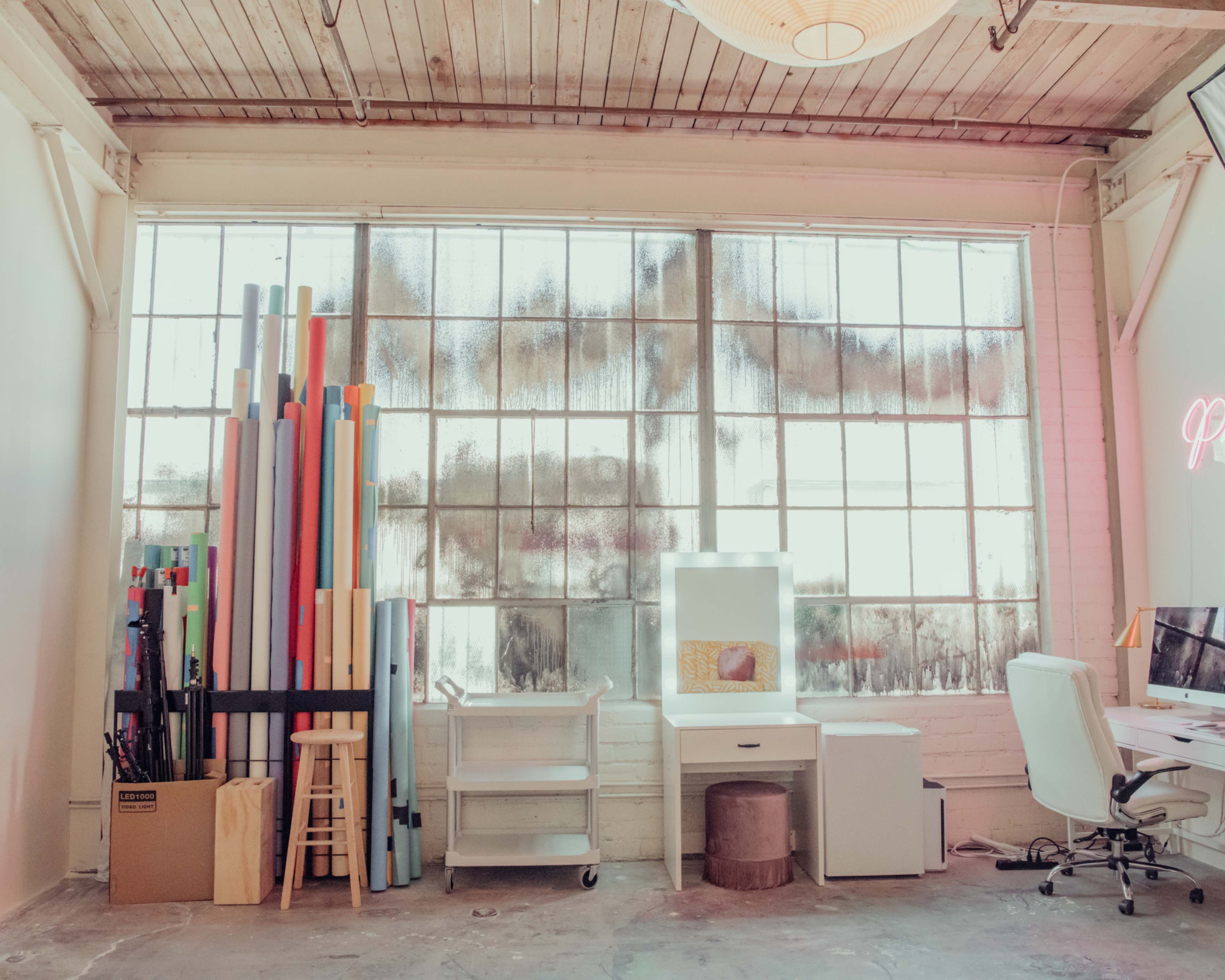 The image shows a workspace with a collection of colorful paper rolls stacked against a large, dusty window, alongside a desk area with a computer and chair.