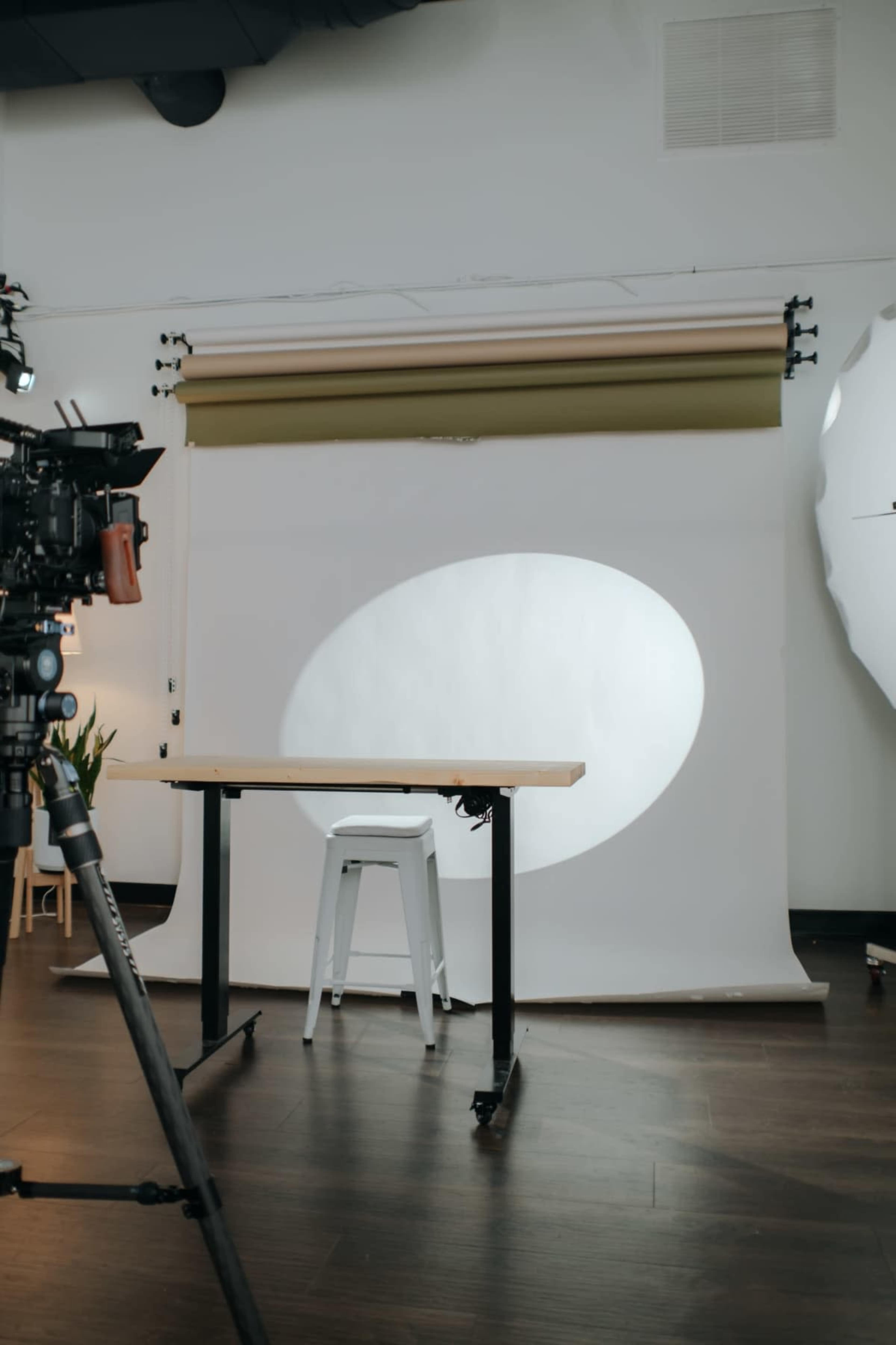 The image shows a photography studio setup with a blank backdrop and a table, accompanied by a camera on a tripod.