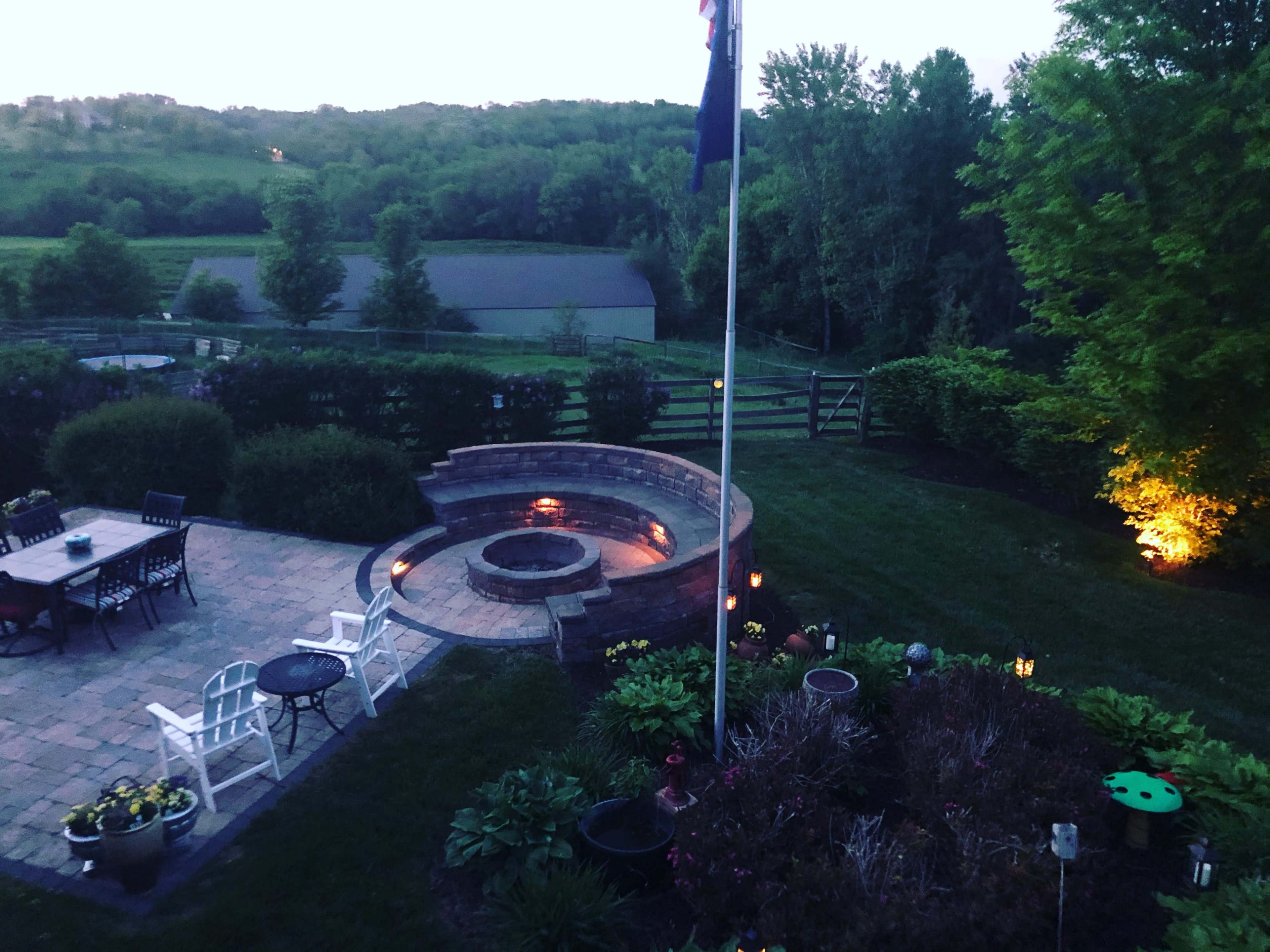 A backyard with a stone fire pit surrounded by seating, a dining area with chairs and a table, and landscaped greenery under evening light.