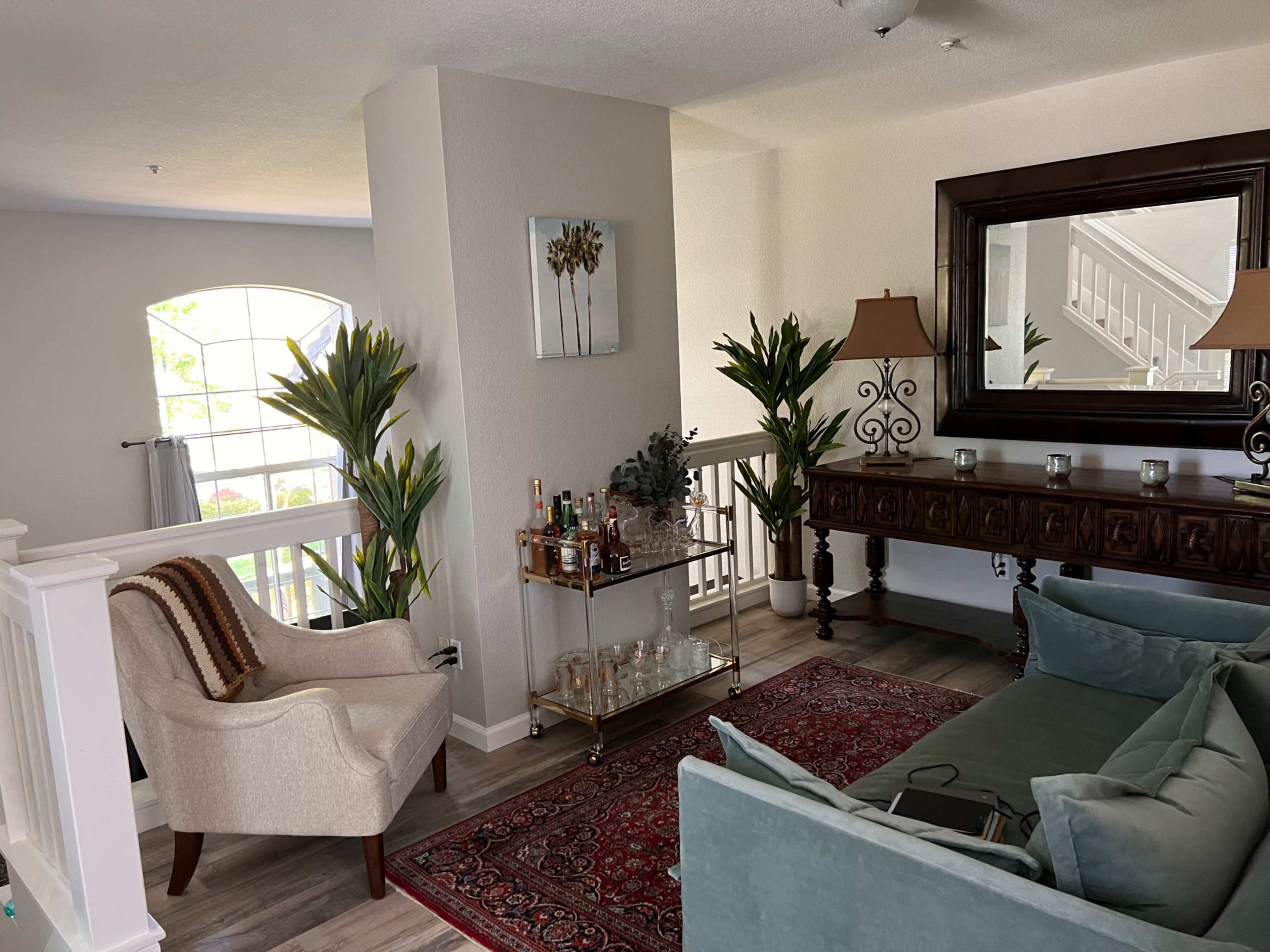 A living room with a sofa, an armchair, a wooden console table with decorative items, and tropical plants, arranged around a small area rug.