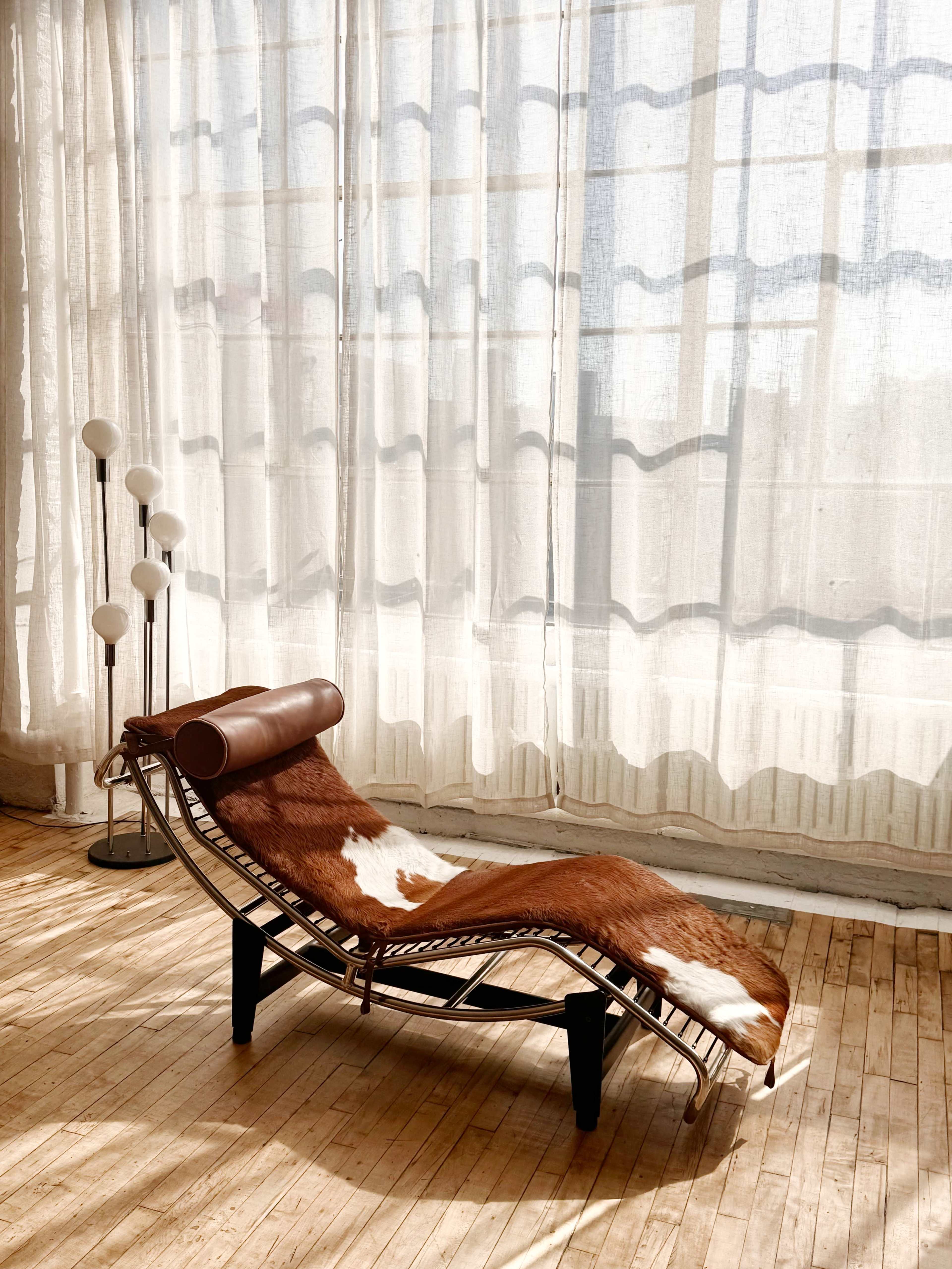A chaise lounge covered with a patterned fabric is positioned near sheer curtains and a floor lamp in a well-lit room.