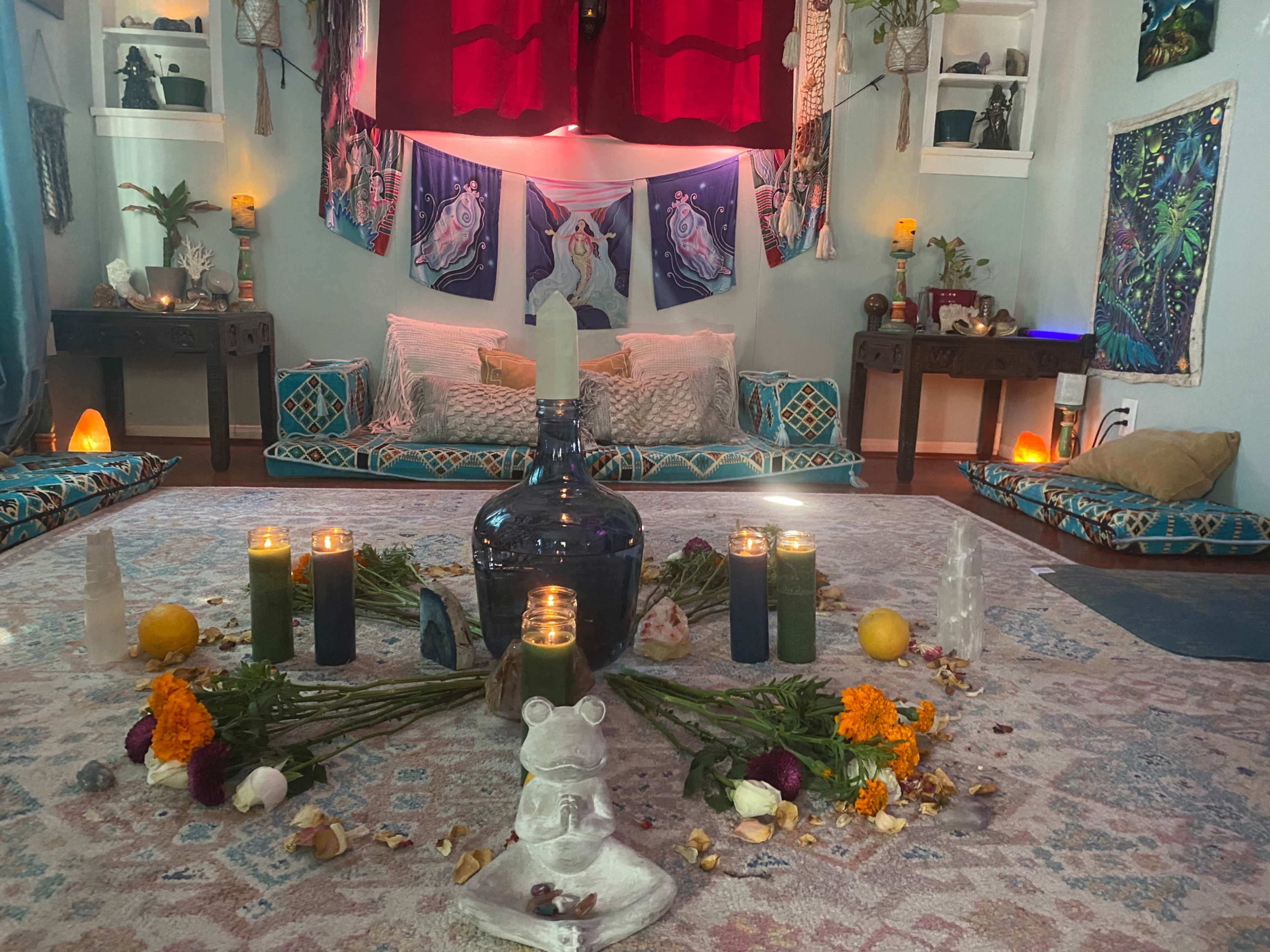 A cozy, well-decorated yoga or meditation space with cushions, candles, flowers, and an ornate centerpiece on a patterned rug.