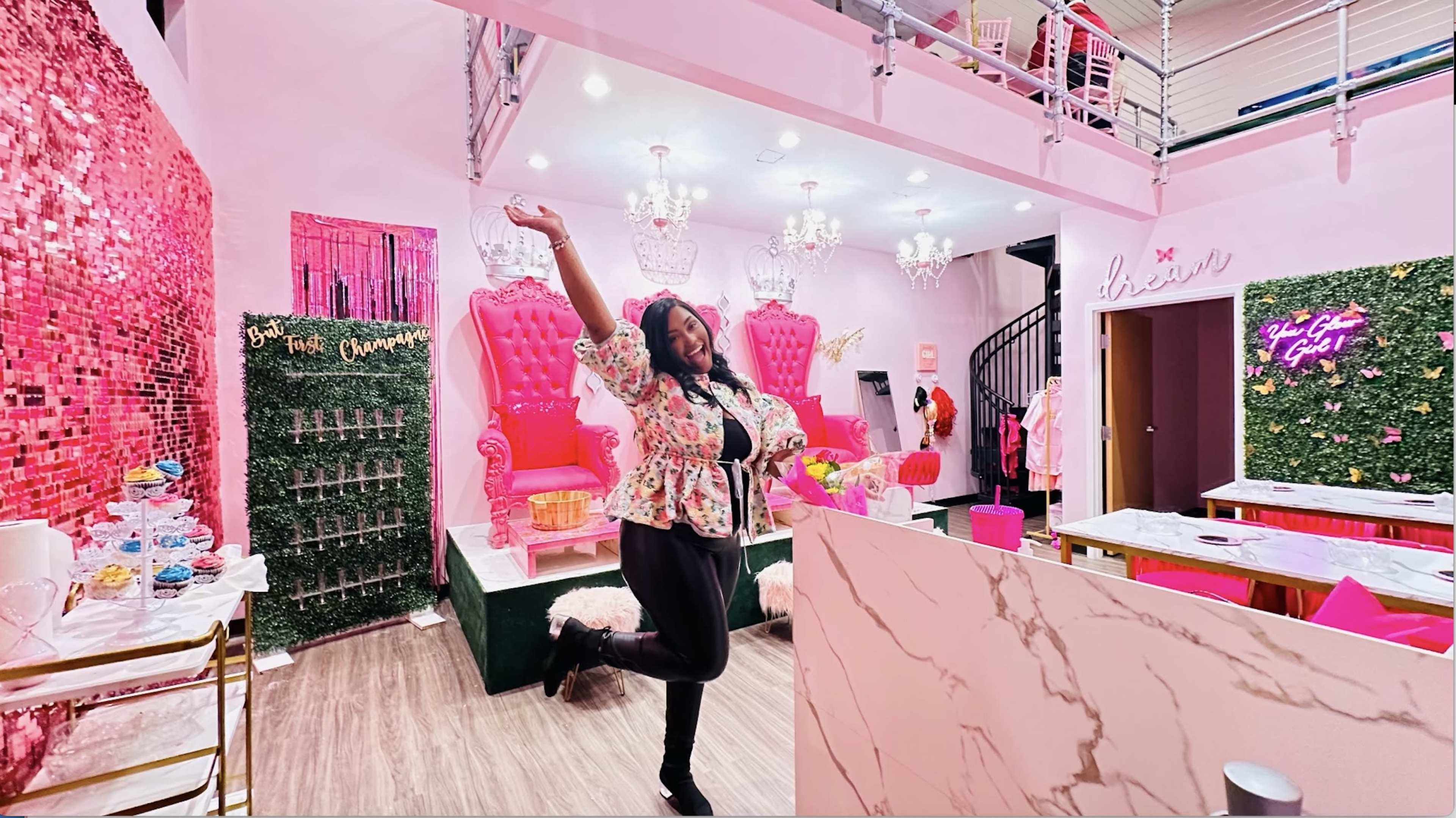 A woman strikes a pose in a brightly decorated room featuring pink chairs, chandeliers, and a marble countertop.