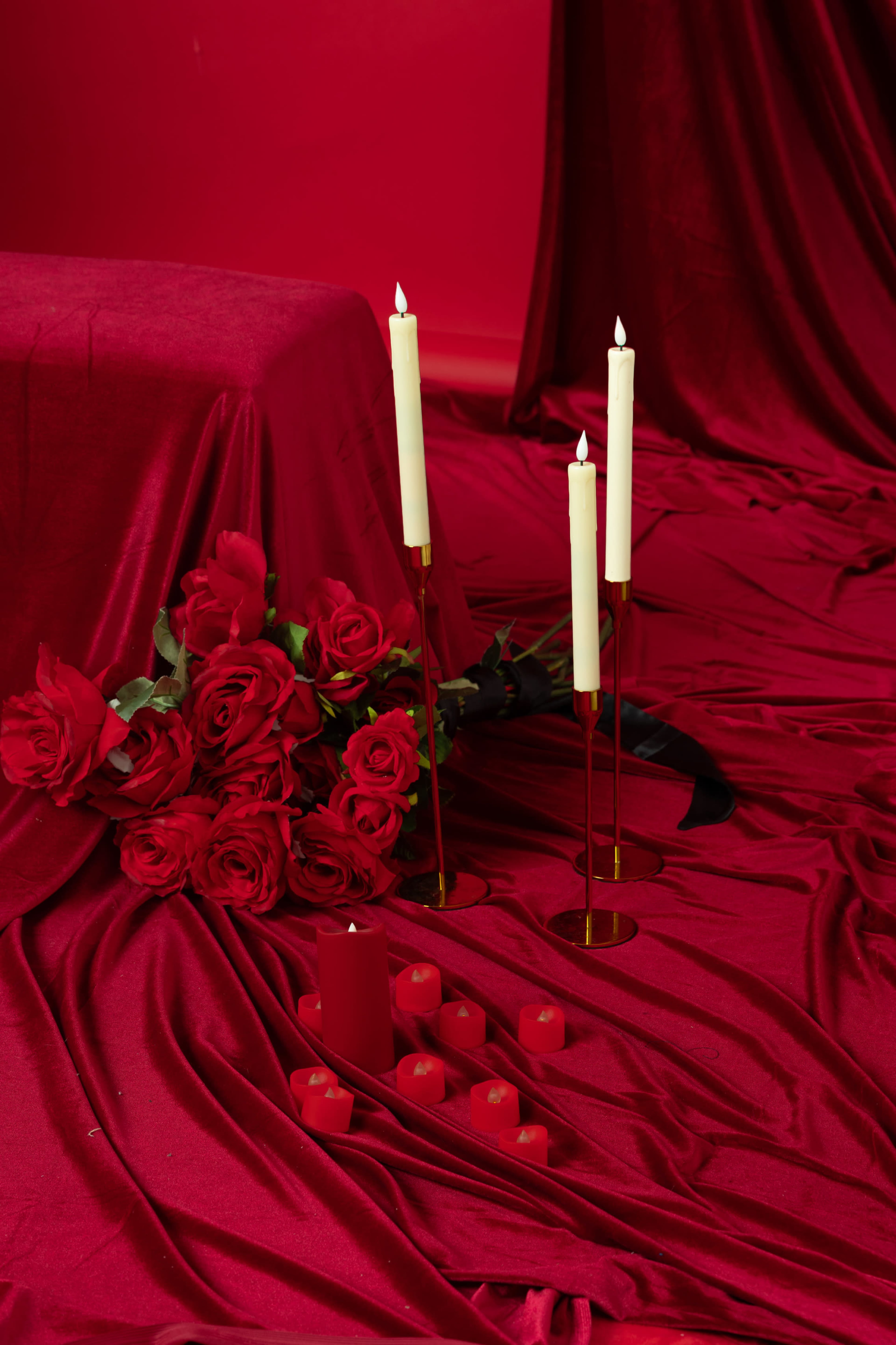 The scene features a vibrant red backdrop with a table covered in red fabric, surrounded by three tall white candles and a cluster of both large and small red candles, alongside a bouquet of red roses.