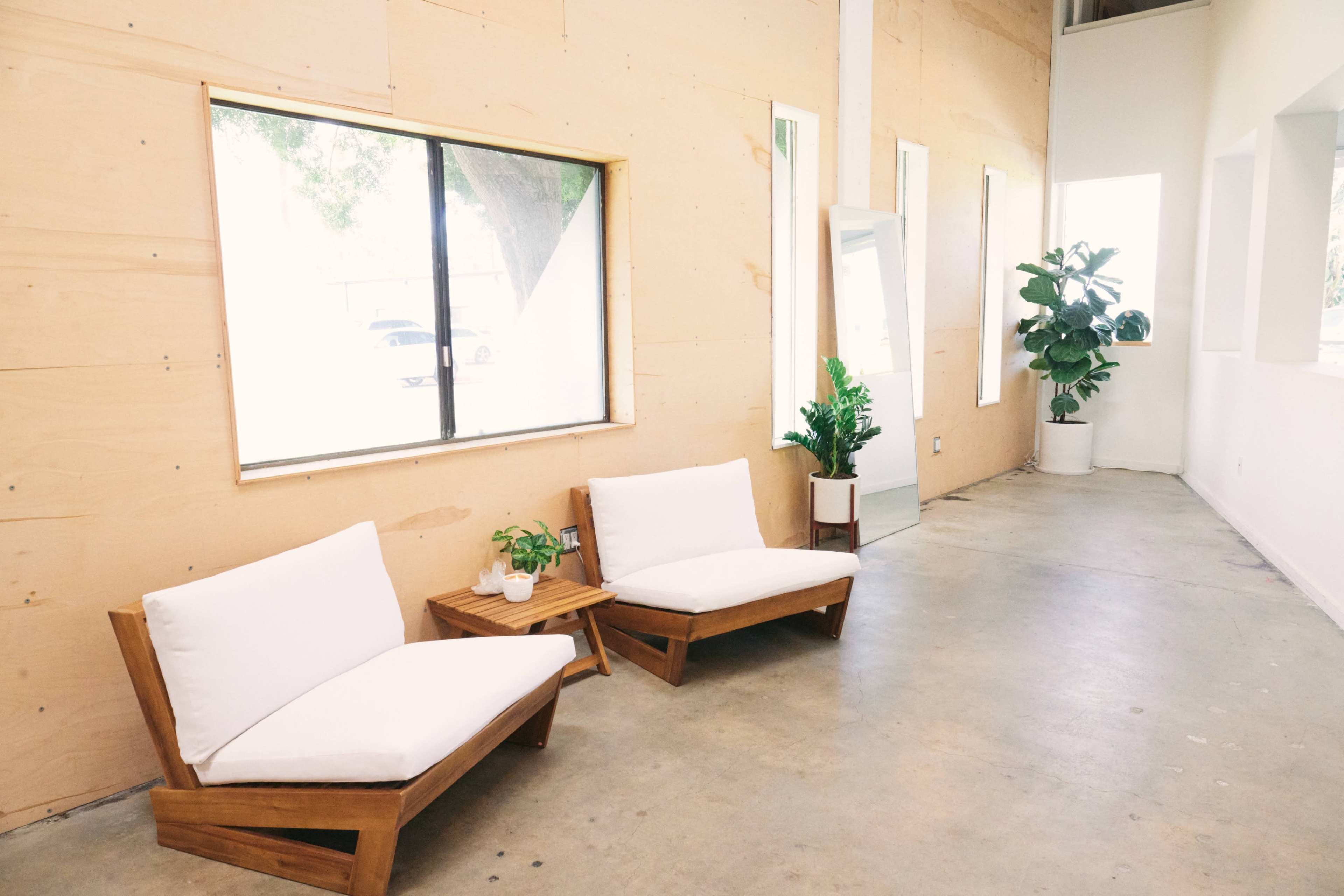 A bright interior space featuring two wooden lounge chairs with white cushions, a small side table with potted plants, and large windows letting in natural light.