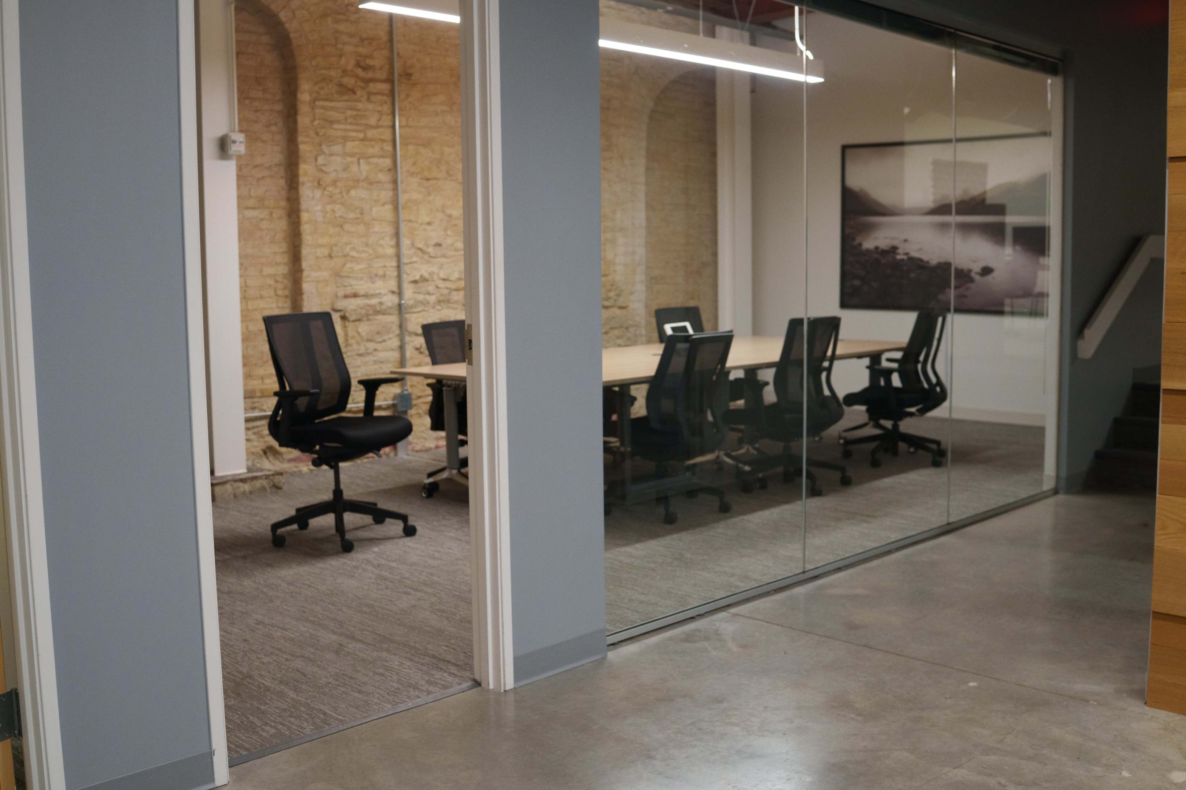 The image shows a modern office space with a glass-walled conference room featuring several black office chairs and a large table, alongside a hallway leading to the room.