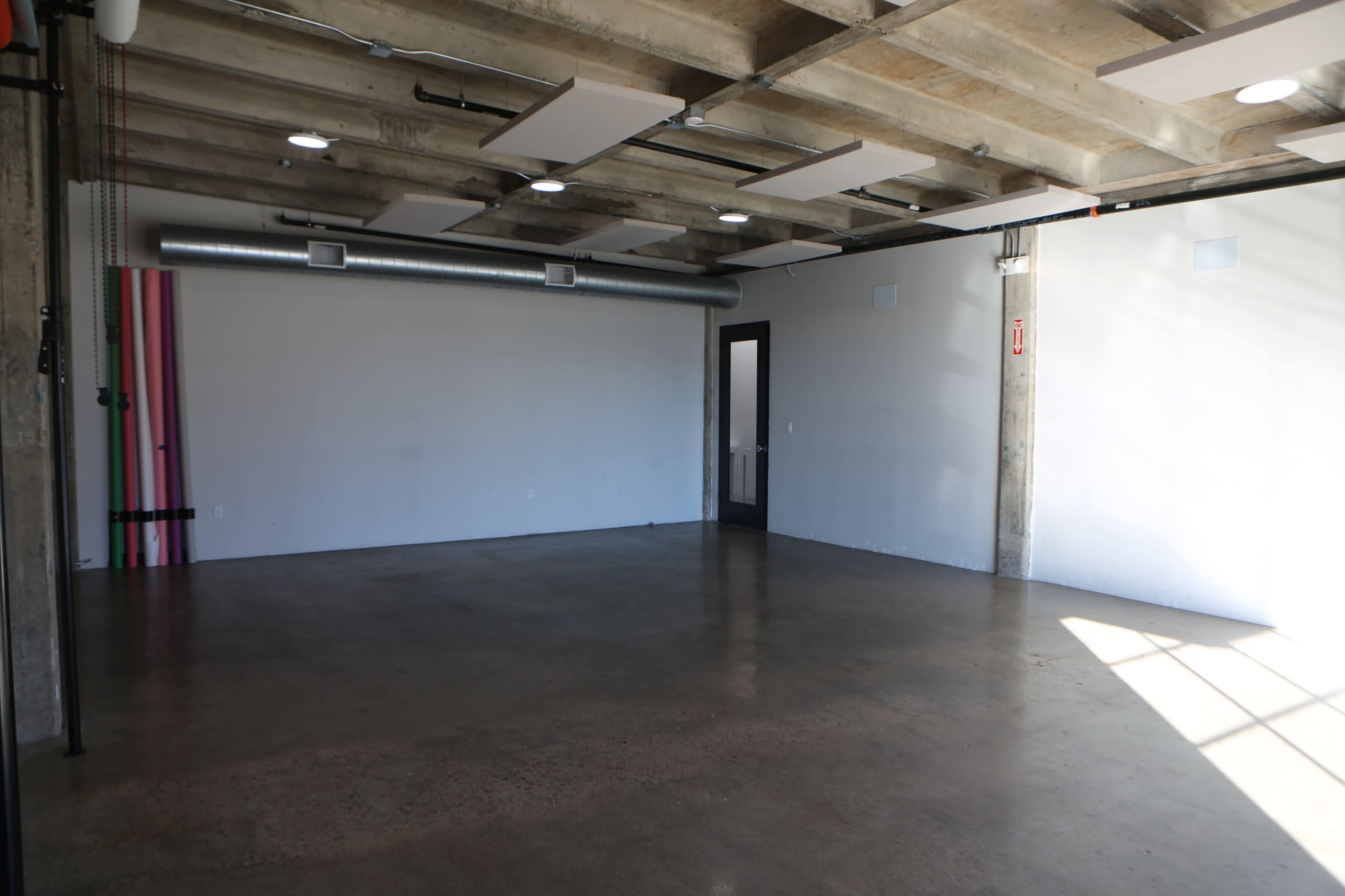 The image shows an empty room with concrete floors, exposed ceiling beams, and a single door on one side.