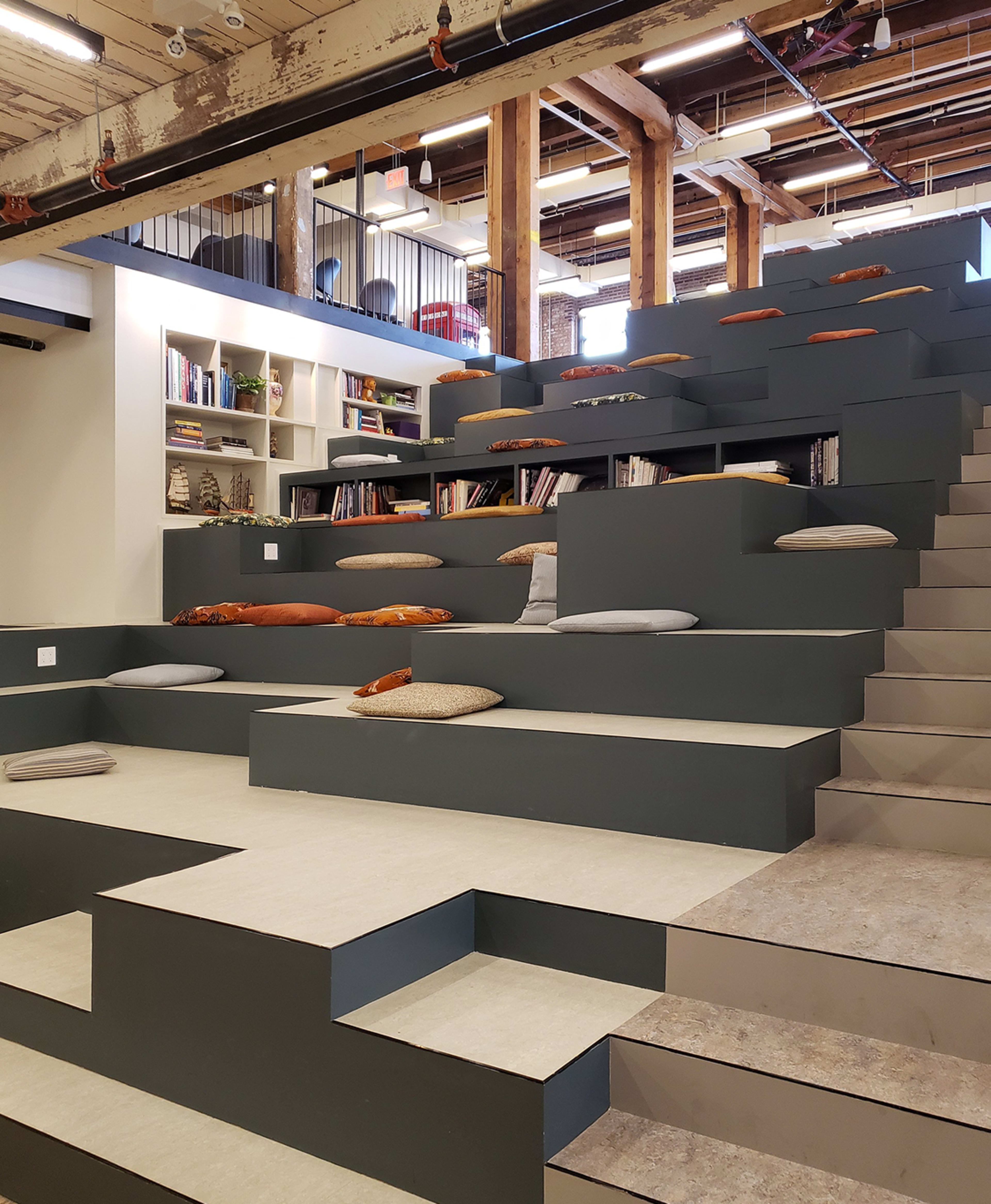 The image shows a multi-level seating area with gray steps and cushions, leading up to a wall lined with shelves filled with books.
