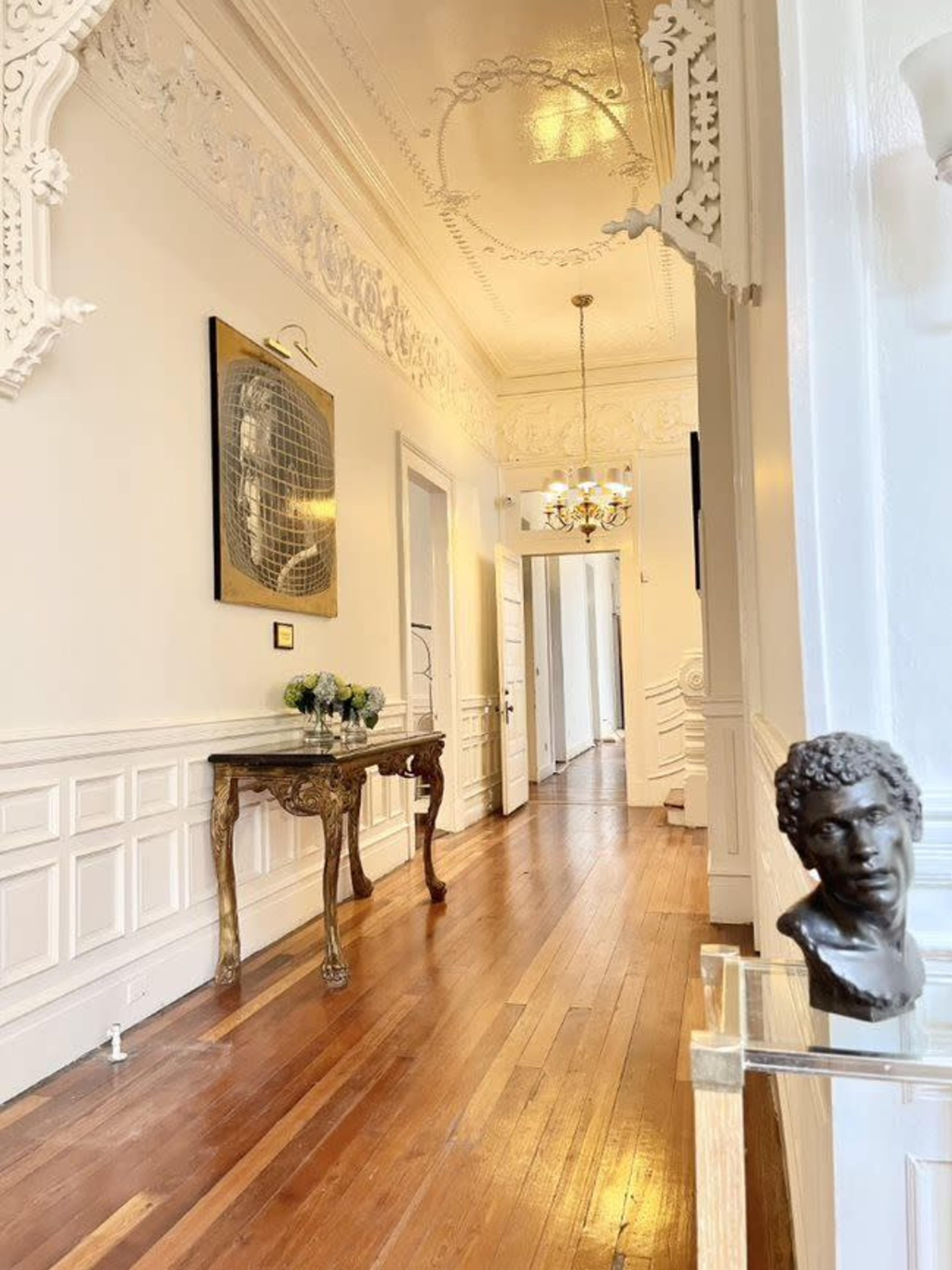 The image shows a long, elegantly designed hallway with hardwood floors, white walls adorned with intricate molding, and a decorative table displaying a bust.