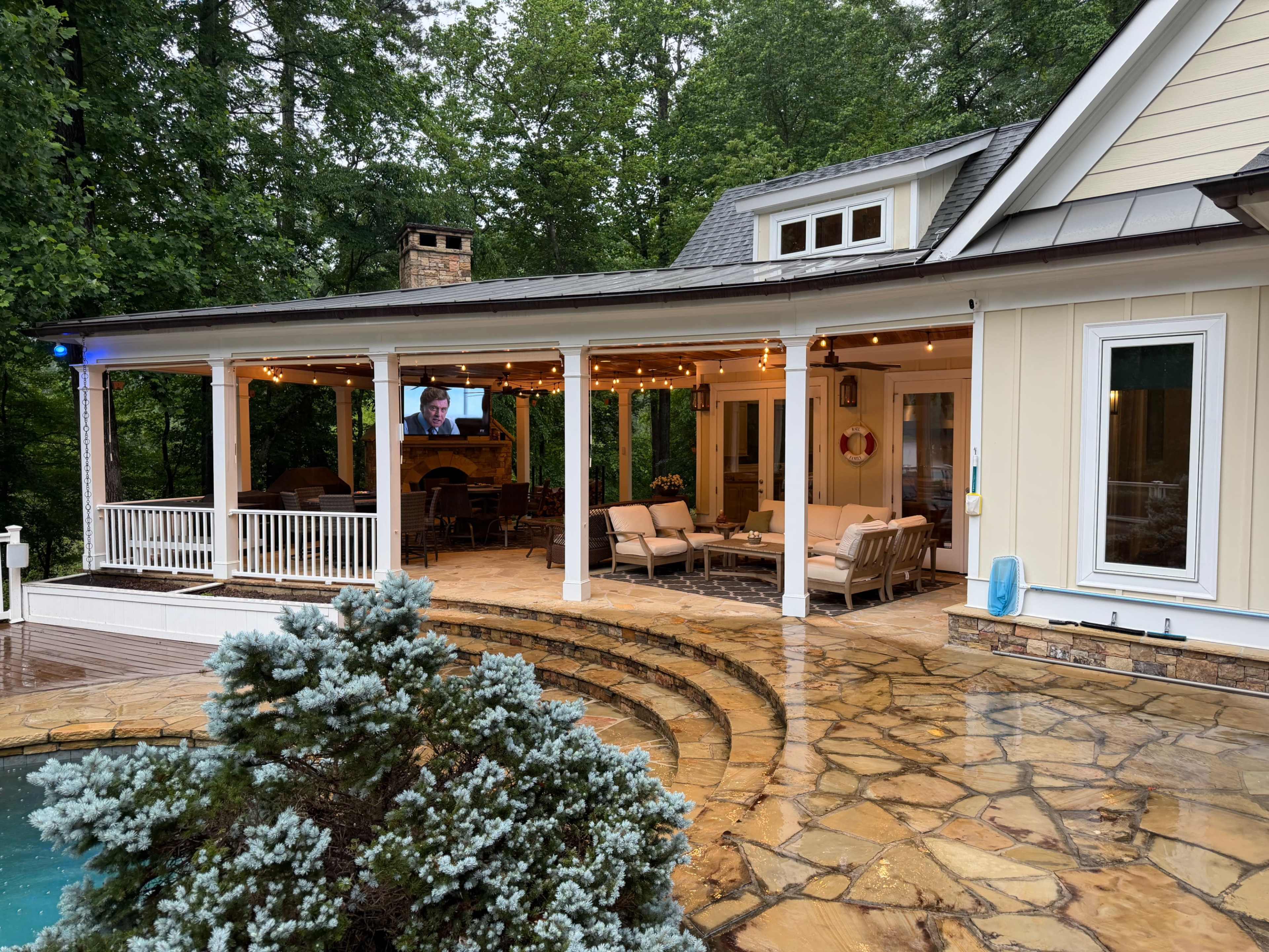 The image shows a porch with several seating arrangements, surrounded by greenery, and featuring a television mounted on the wall.