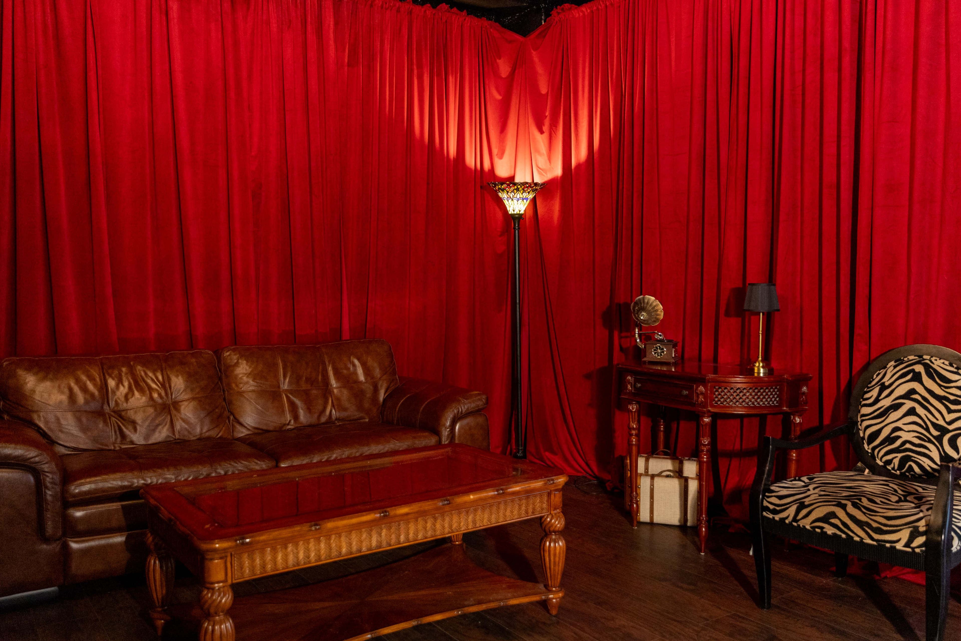 The scene features a cozy living area with a brown leather sofa, a wooden coffee table, and a zebra-striped armchair, surrounded by red drapes and warm lighting.
