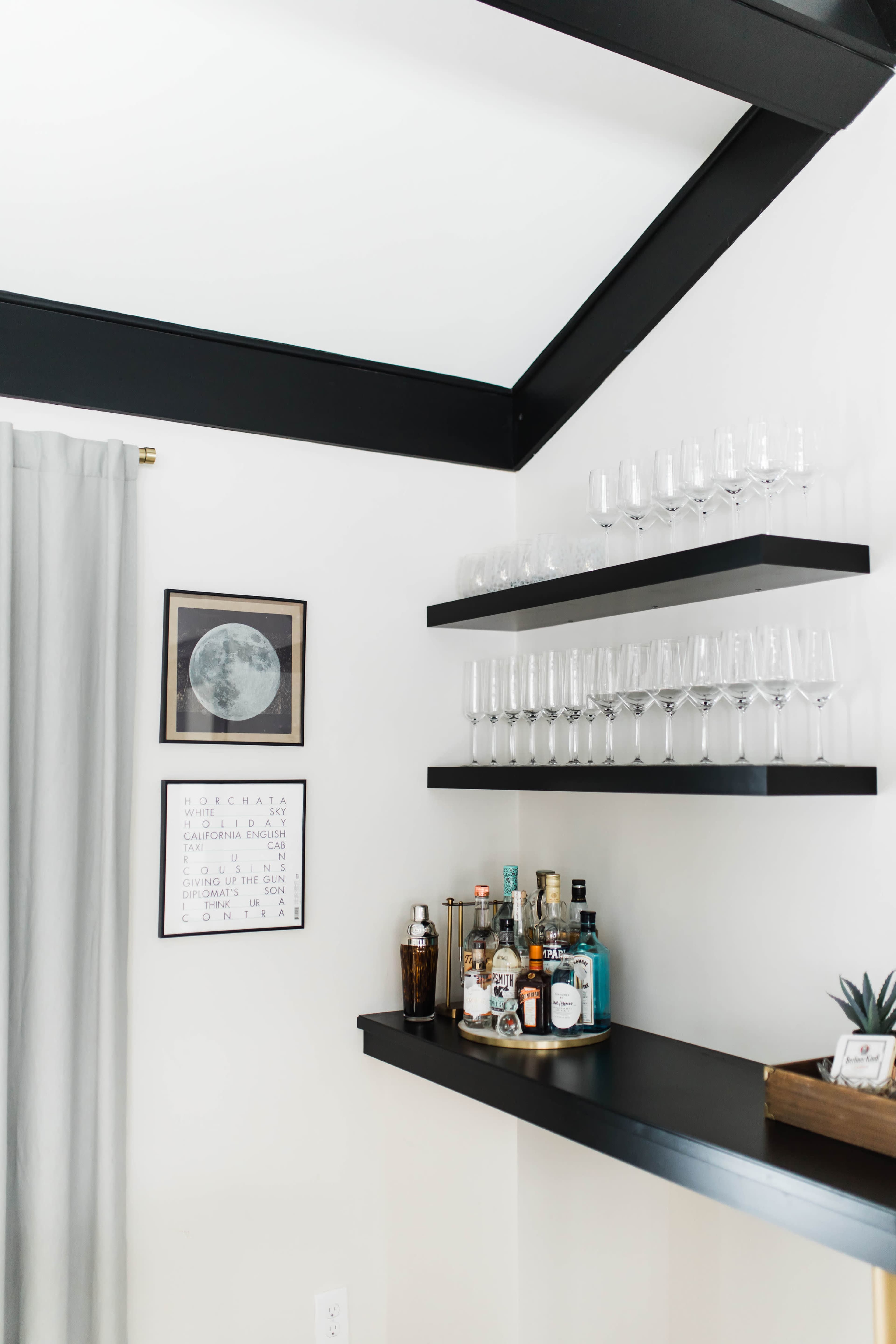 A black wooden shelf holds several liquor bottles and glasses against a white wall, with a framed moon artwork and a decorative print nearby.
