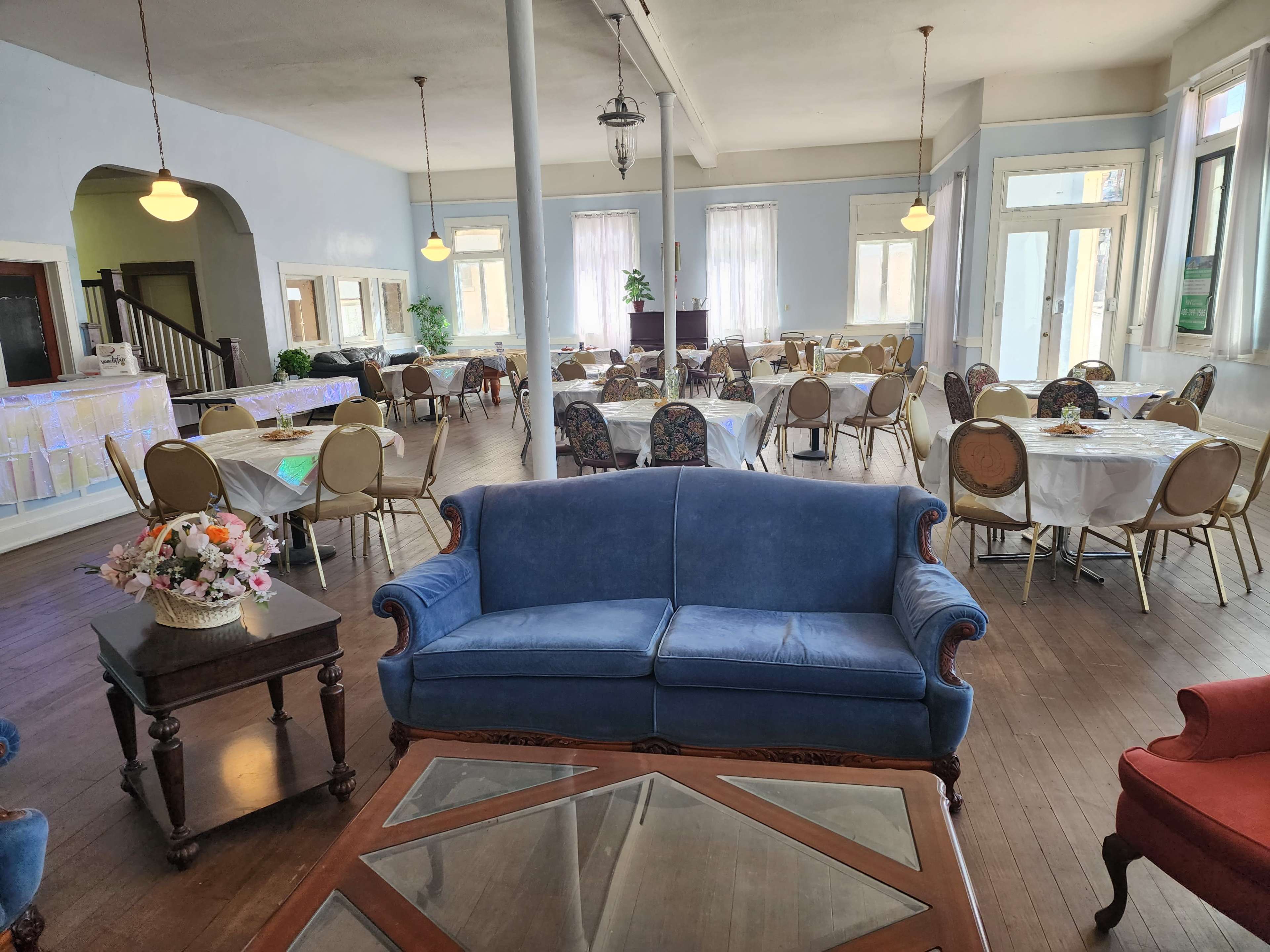 A spacious dining area with tables set for meals, a blue sofa in the foreground, and large windows allowing natural light to enter.