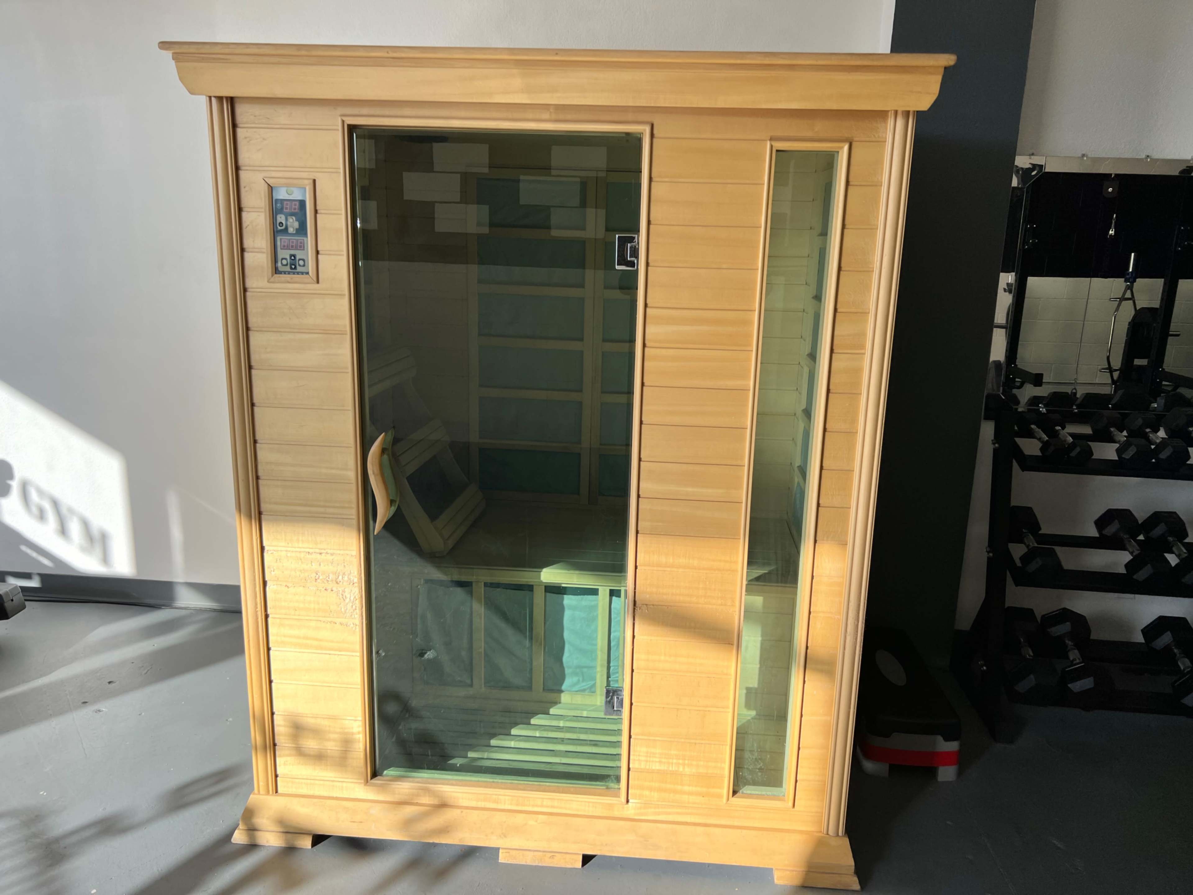 A wooden sauna with a glass door and control panel, positioned in a gym setting.