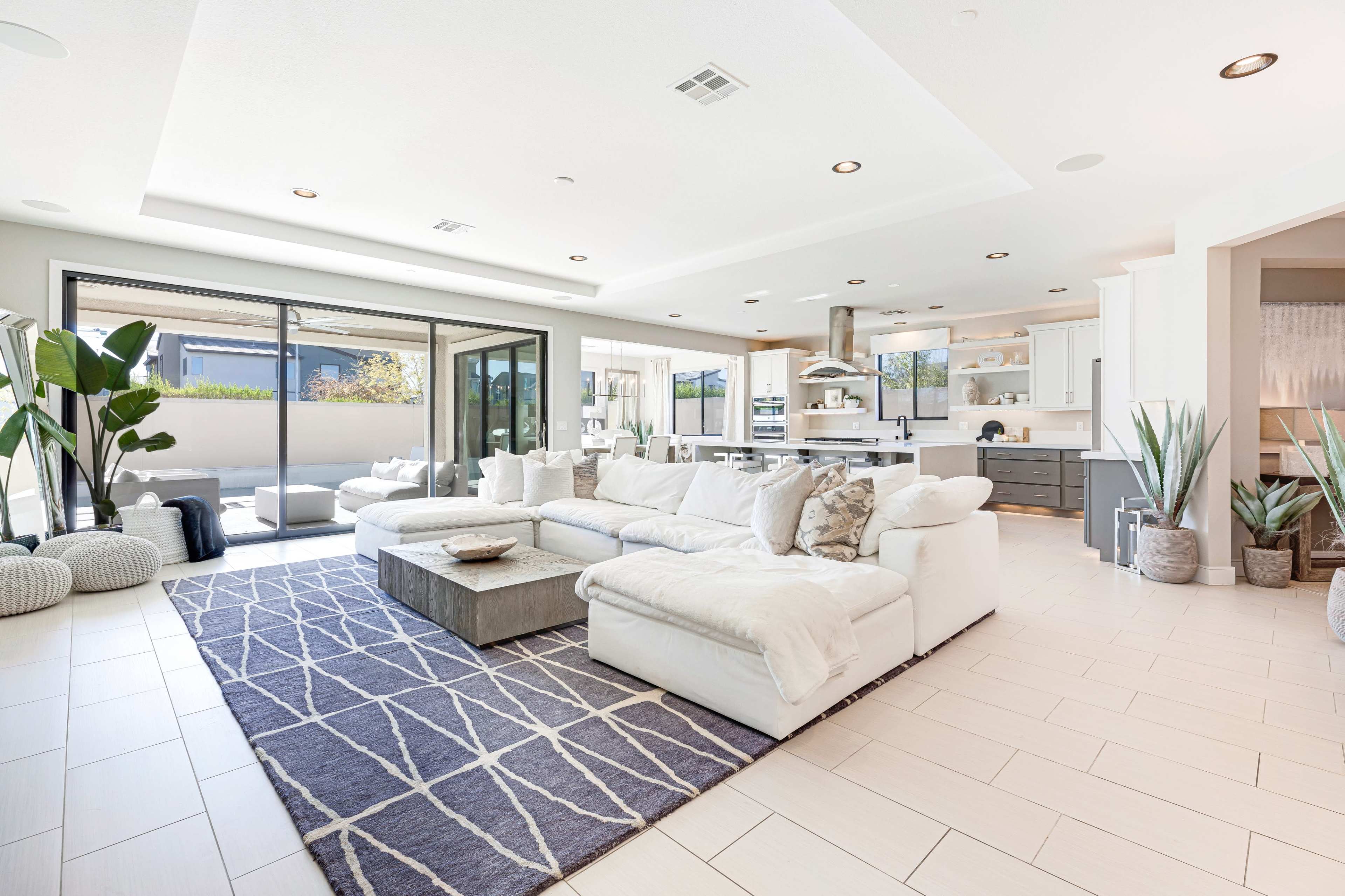 A spacious living room features a large white sectional sofa, a wooden coffee table, and an open layout leading to a well-lit kitchen.