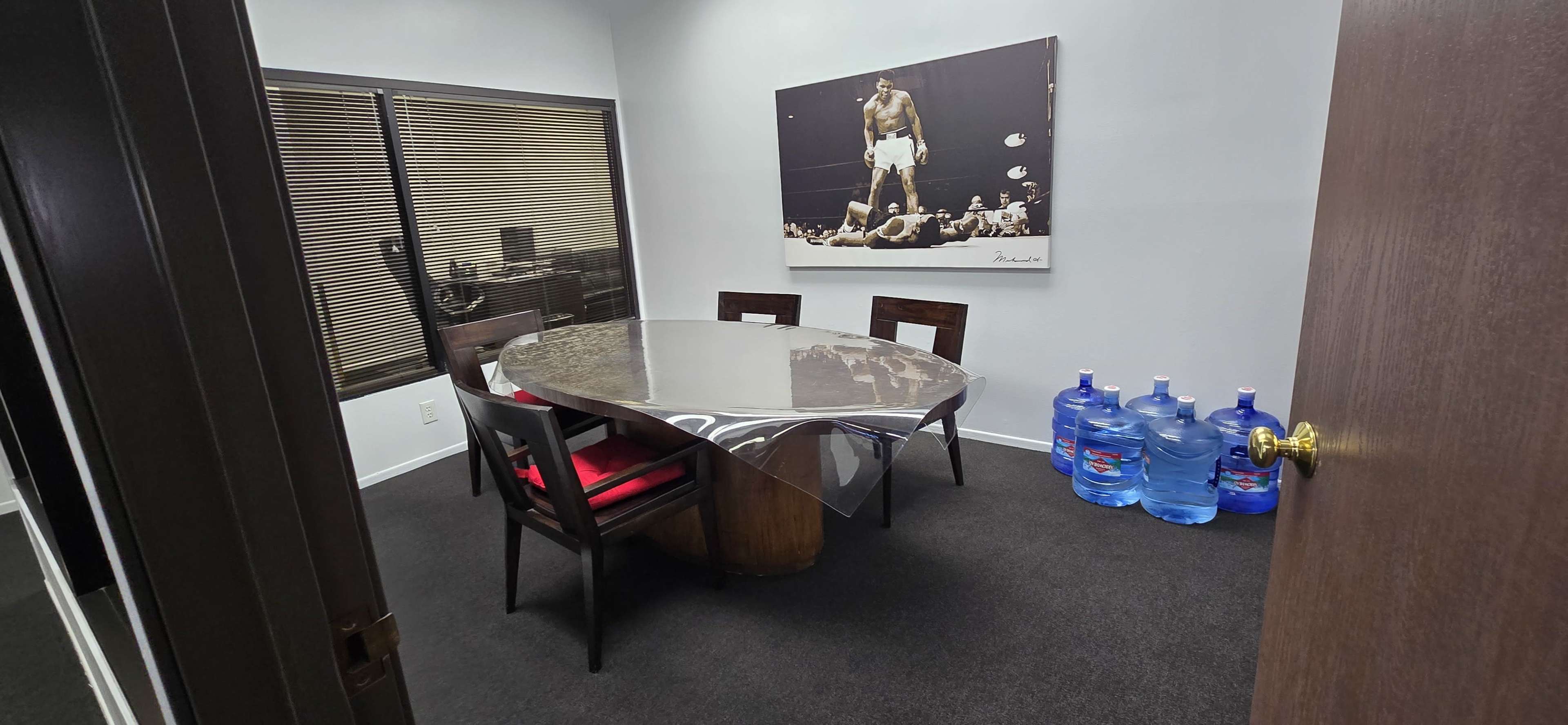 A conference room featuring a round table with a metallic surface, surrounded by wooden chairs, and a large photo on the wall of a boxer, alongside several water jugs stacked on the floor.