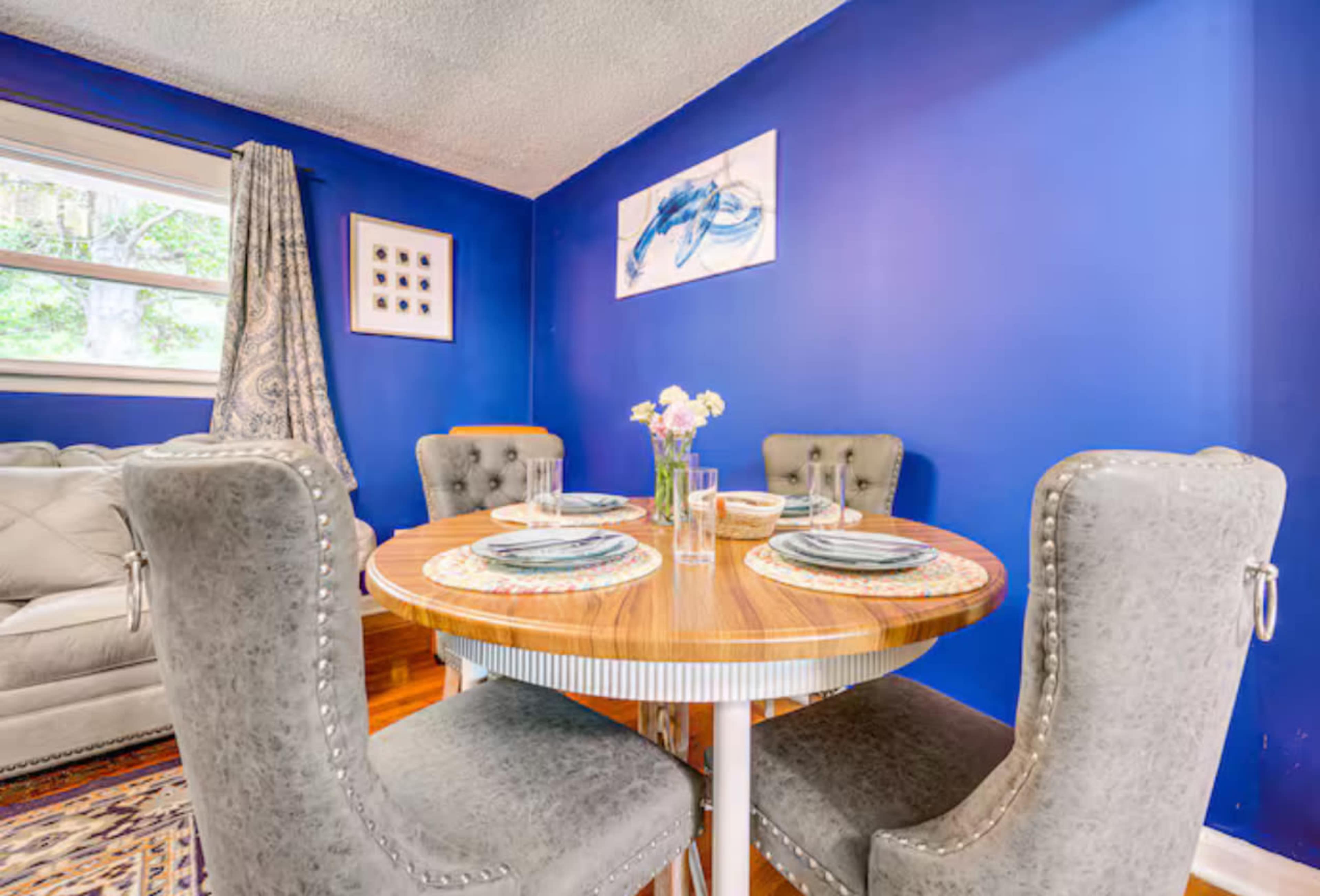 A round dining table is set with plates and utensils, surrounded by four tufted chairs in a blue-walled room that includes a gray couch and a floral centerpiece.