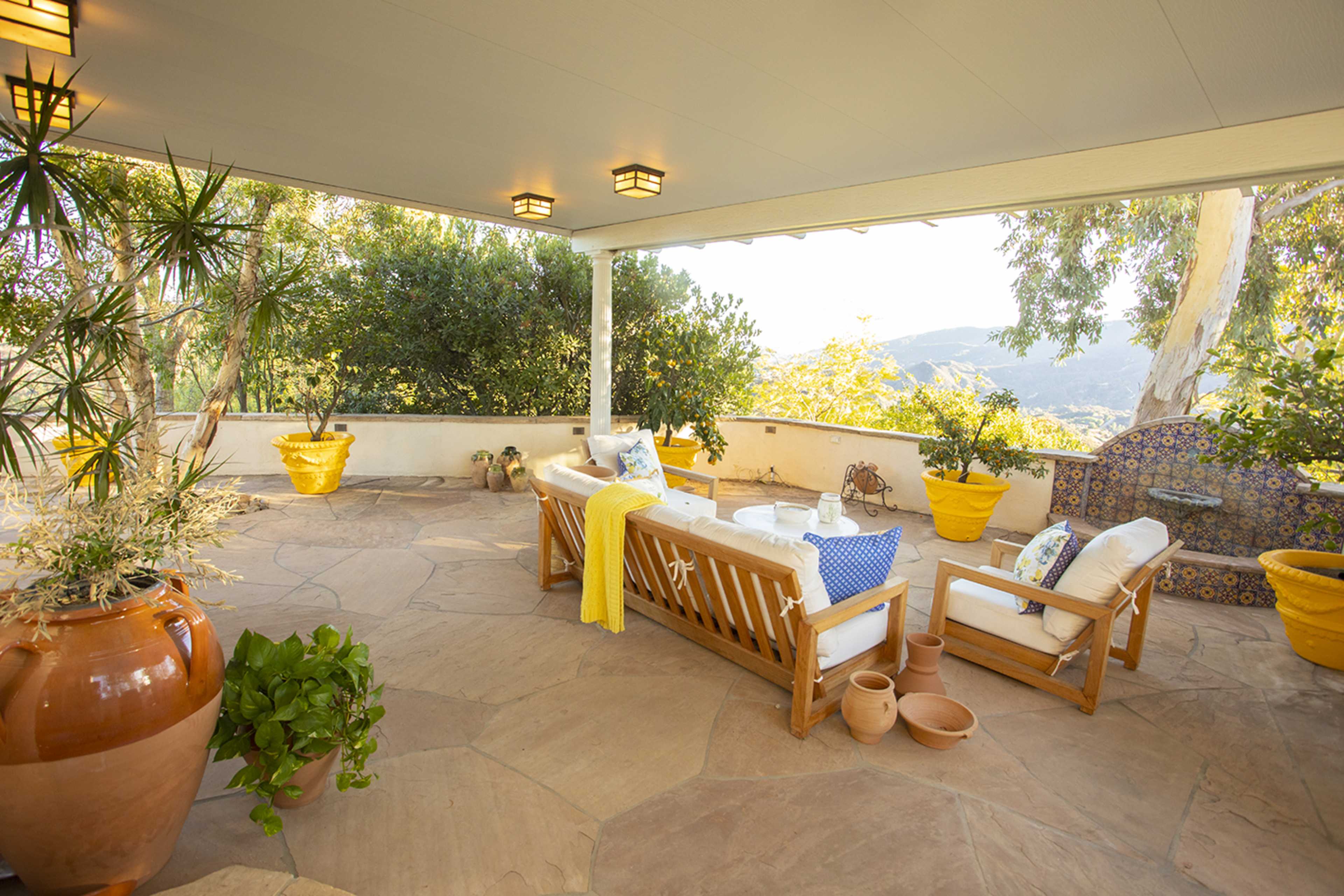 A cozy outdoor seating area features wooden furniture, potted plants, and vibrant yellow planters, with a view of the surrounding landscape.