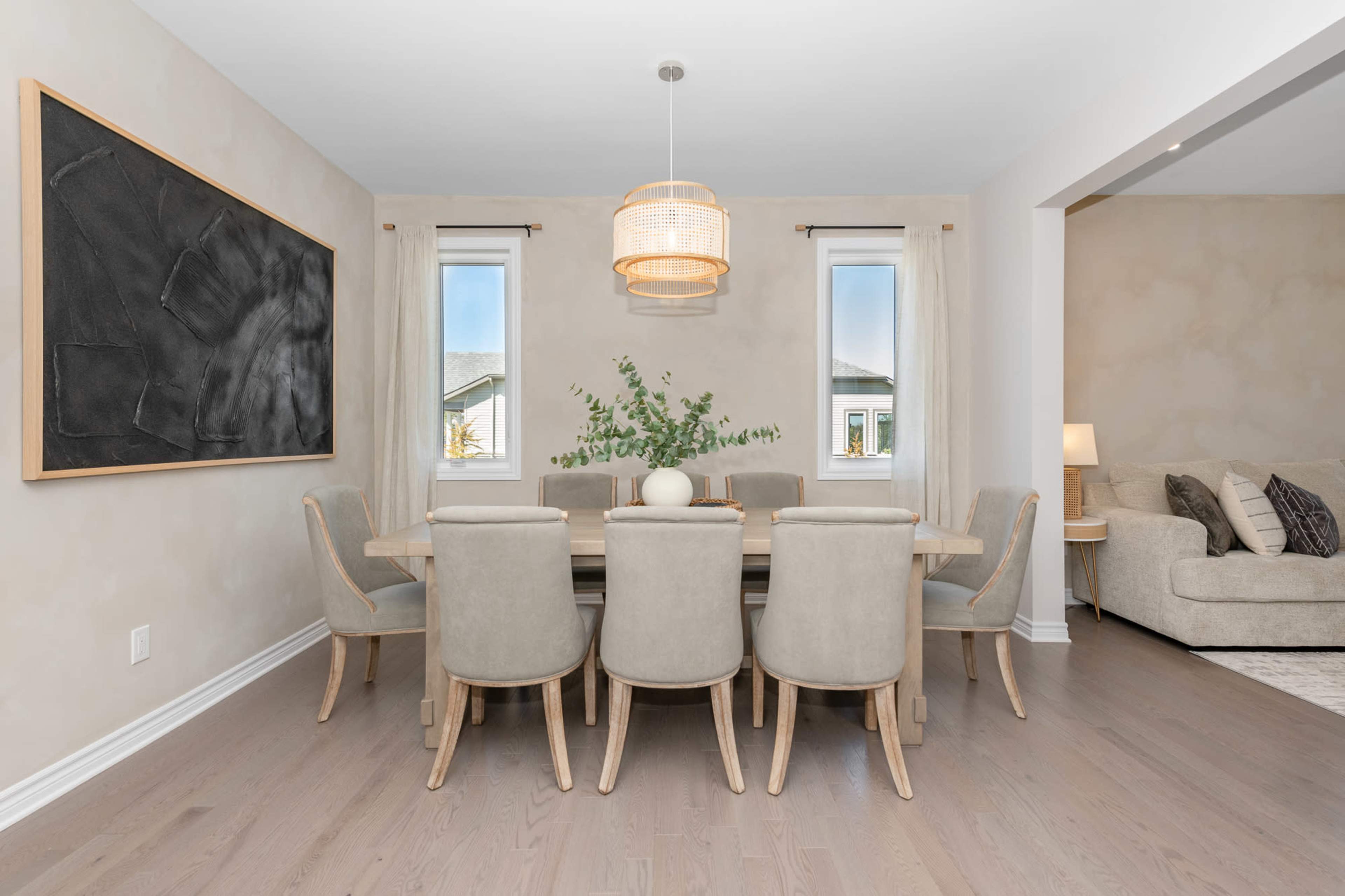 A modern dining room features a large table surrounded by upholstered chairs, with a pendant light above and windows on either side.