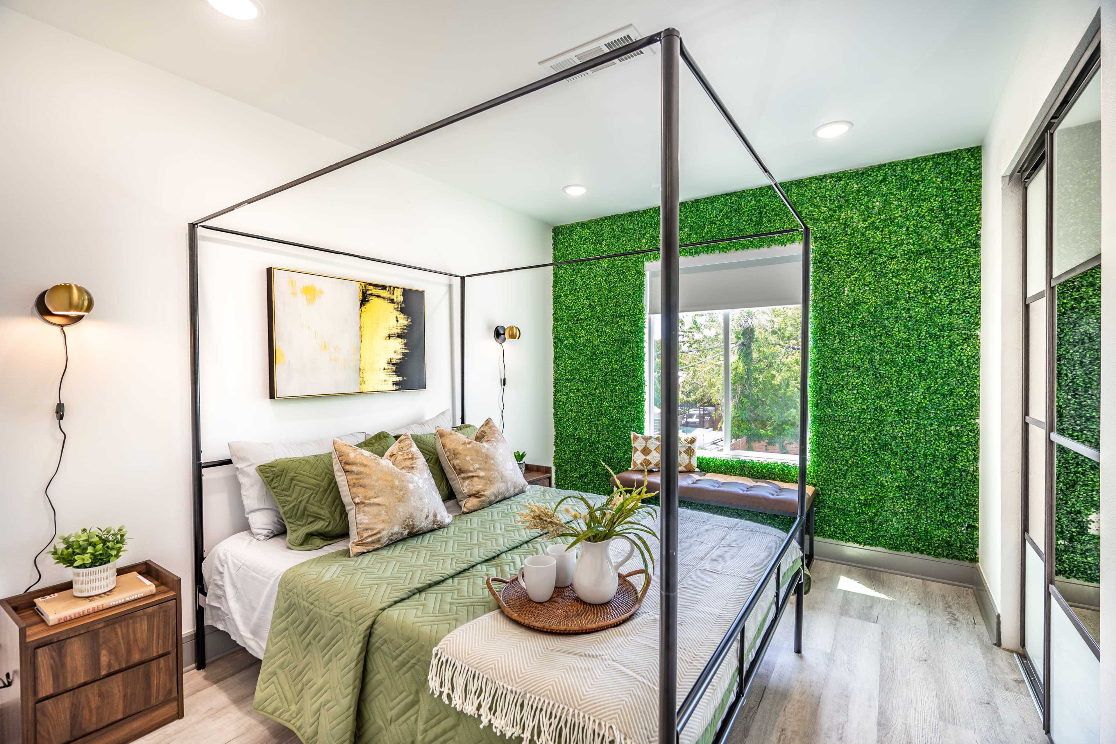 A modern bedroom features a four-poster bed with green accents, complemented by a wall of artificial grass and large windows.