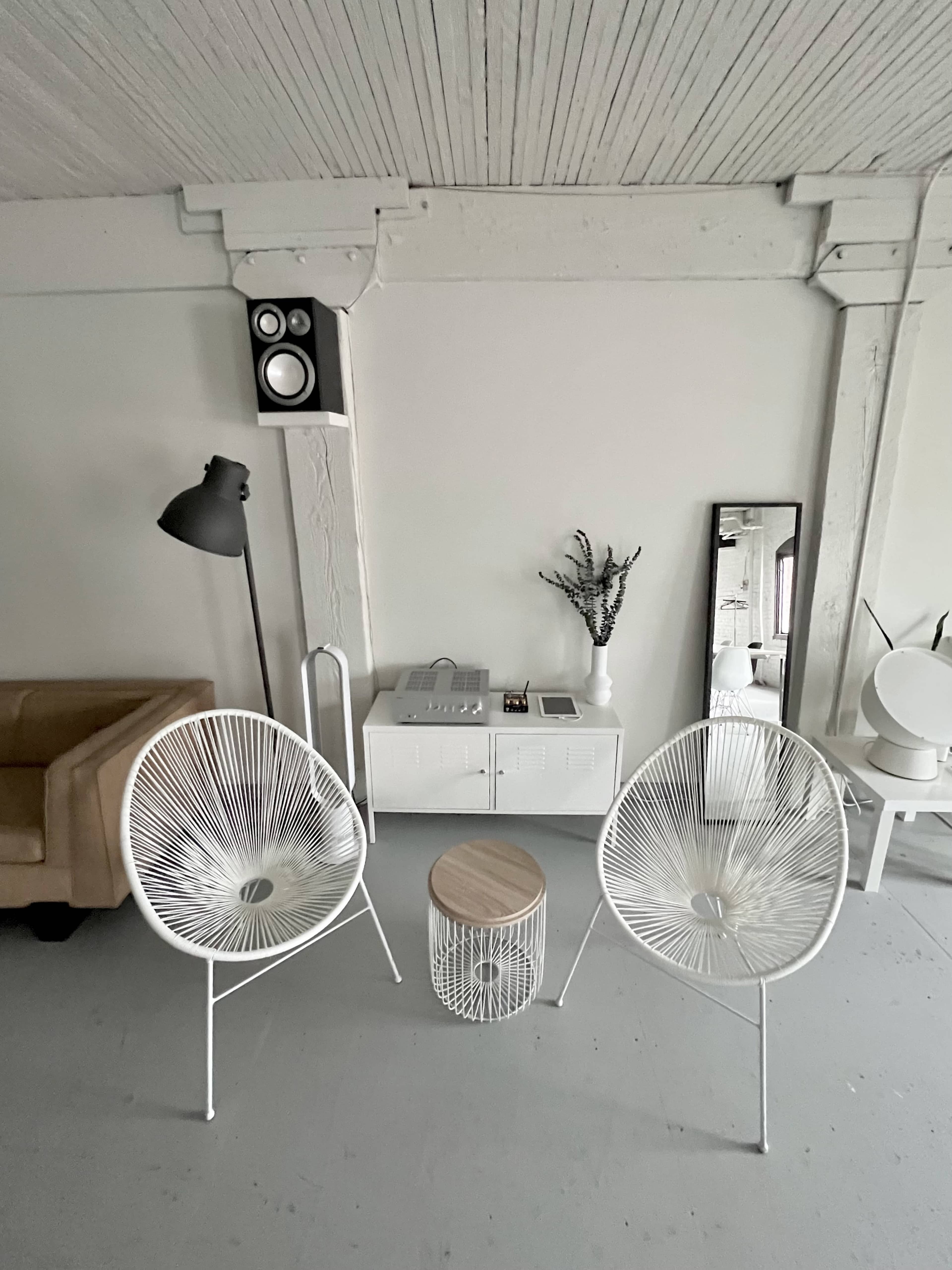 A minimalistic interior featuring two white chairs, a small wooden table, a floor lamp, a wall-mounted speaker, and a console table with a mirror and decorative plant.
