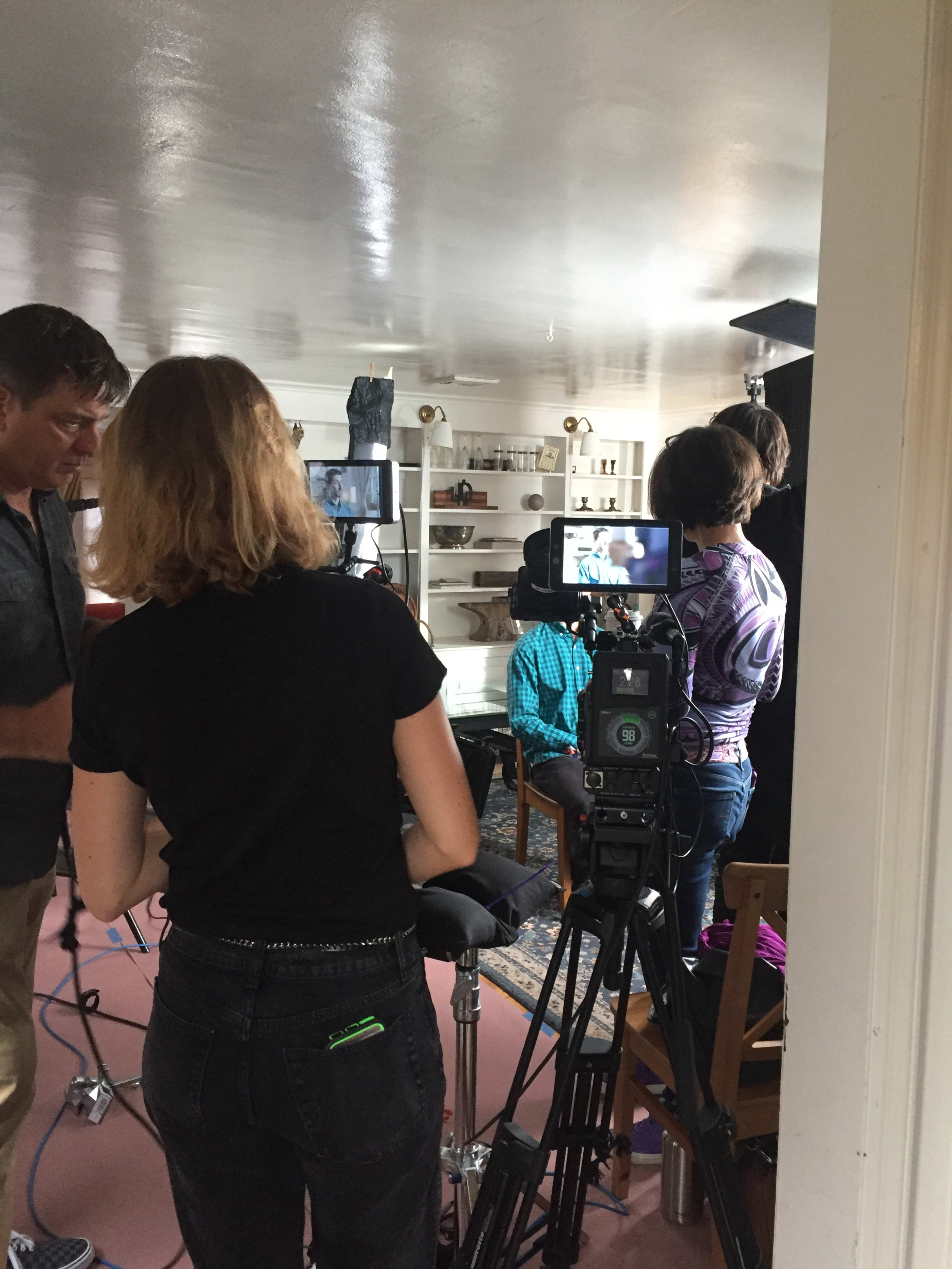 A film crew sets up equipment in a brightly lit room with a table, preparing for an interview scene.