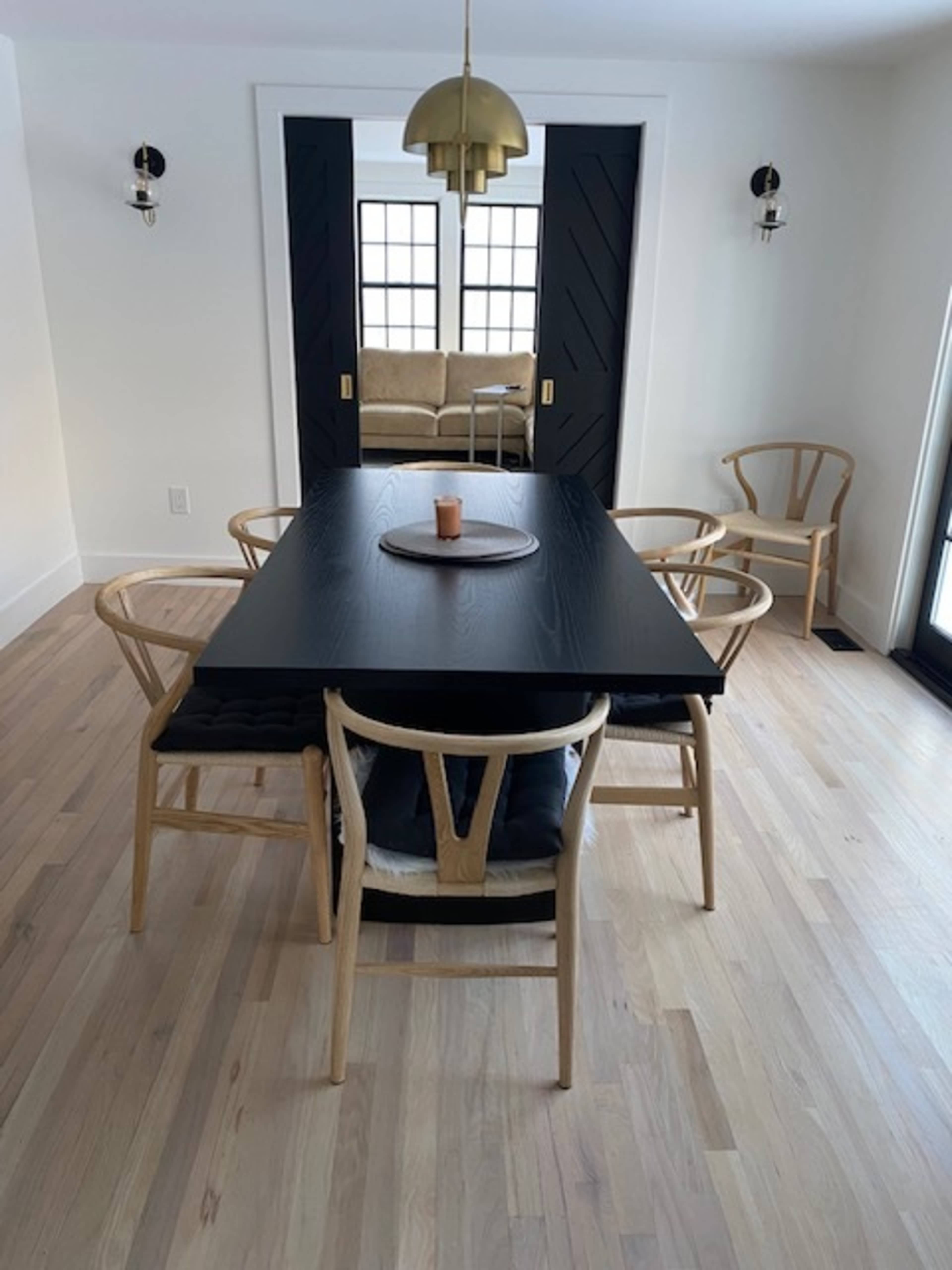 A modern dining area features a long black table surrounded by light wooden chairs, with a pendant light above and a decorative couch visible through double doors.
