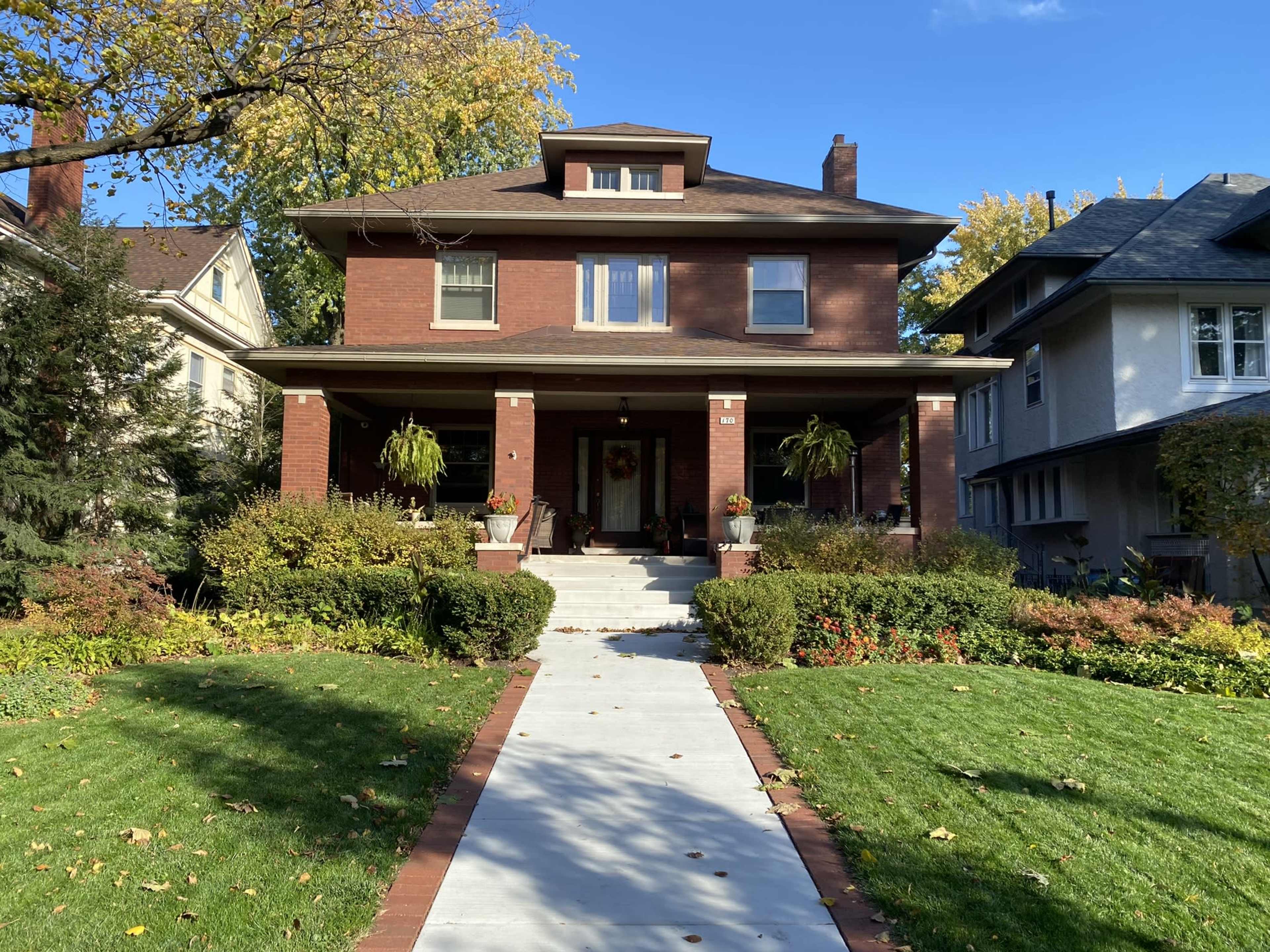 The image shows a two-story brick house with a covered front porch, surrounded by a neatly landscaped yard and a pathway leading to the entrance.