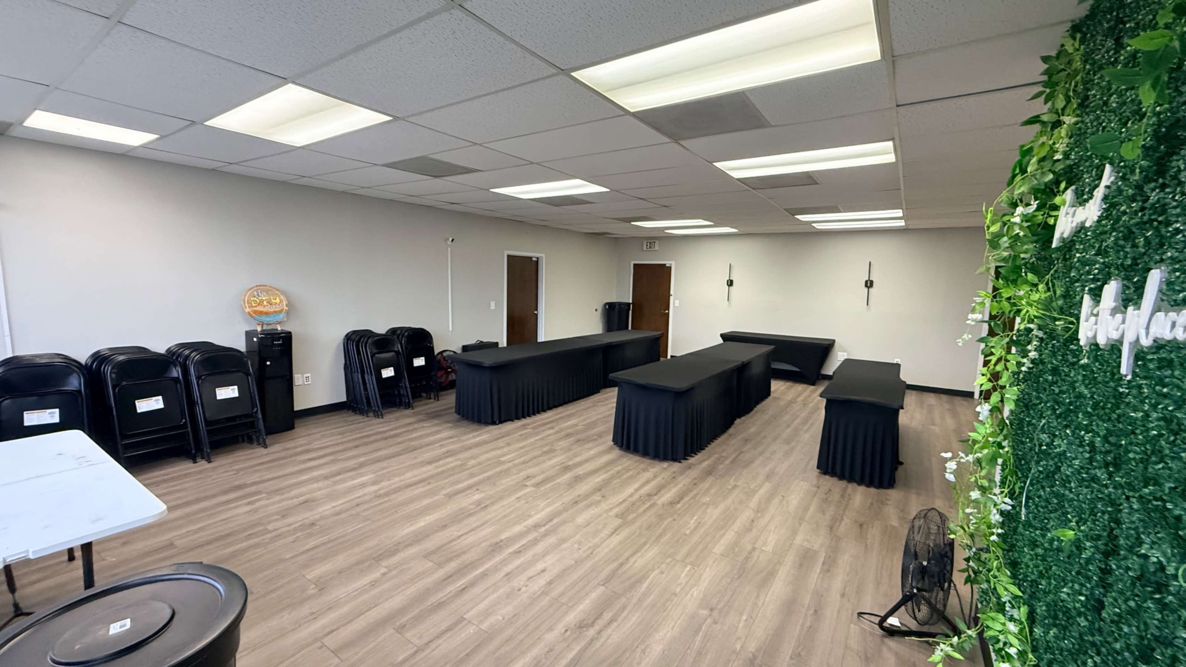 The image shows a mostly empty room with several black folding chairs stacked against one wall and long tables covered with black skirts arranged throughout the space.