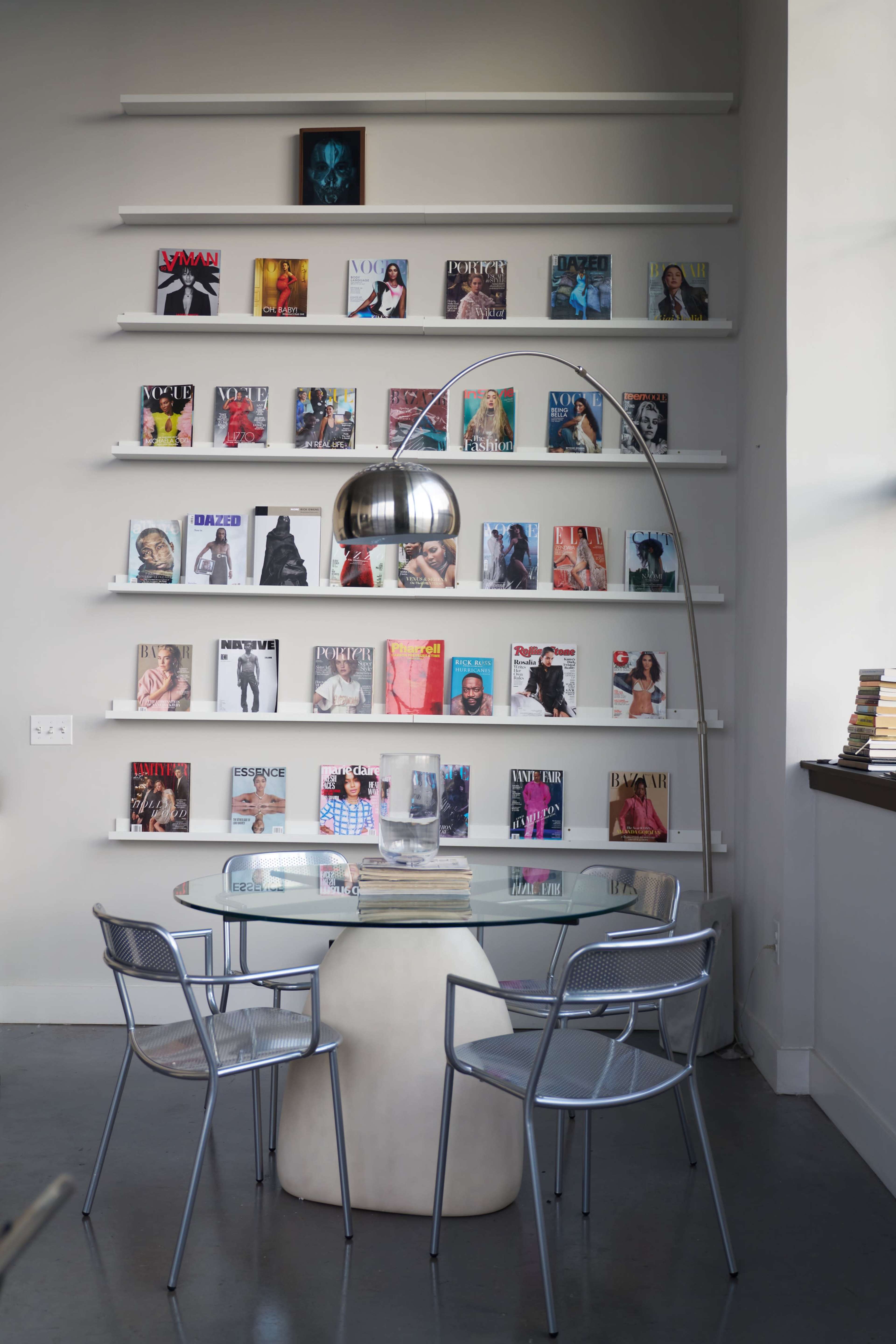A modern reading nook features a glass table with two metal chairs, surrounded by shelves displaying various magazine covers on a gray wall.