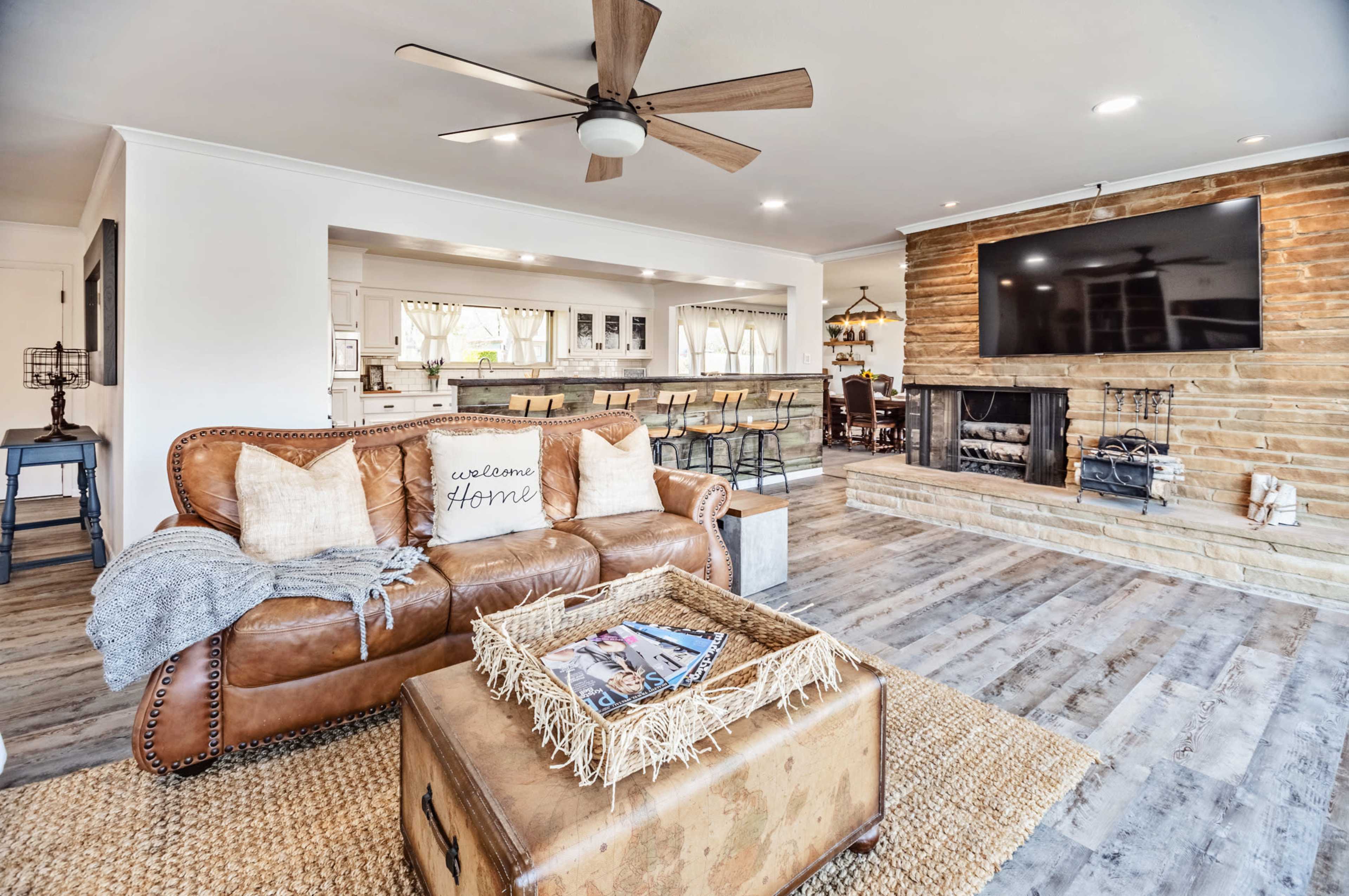 The image shows a living room with a leather sofa, a coffee table, a stone fireplace, and an open kitchen area in the background.