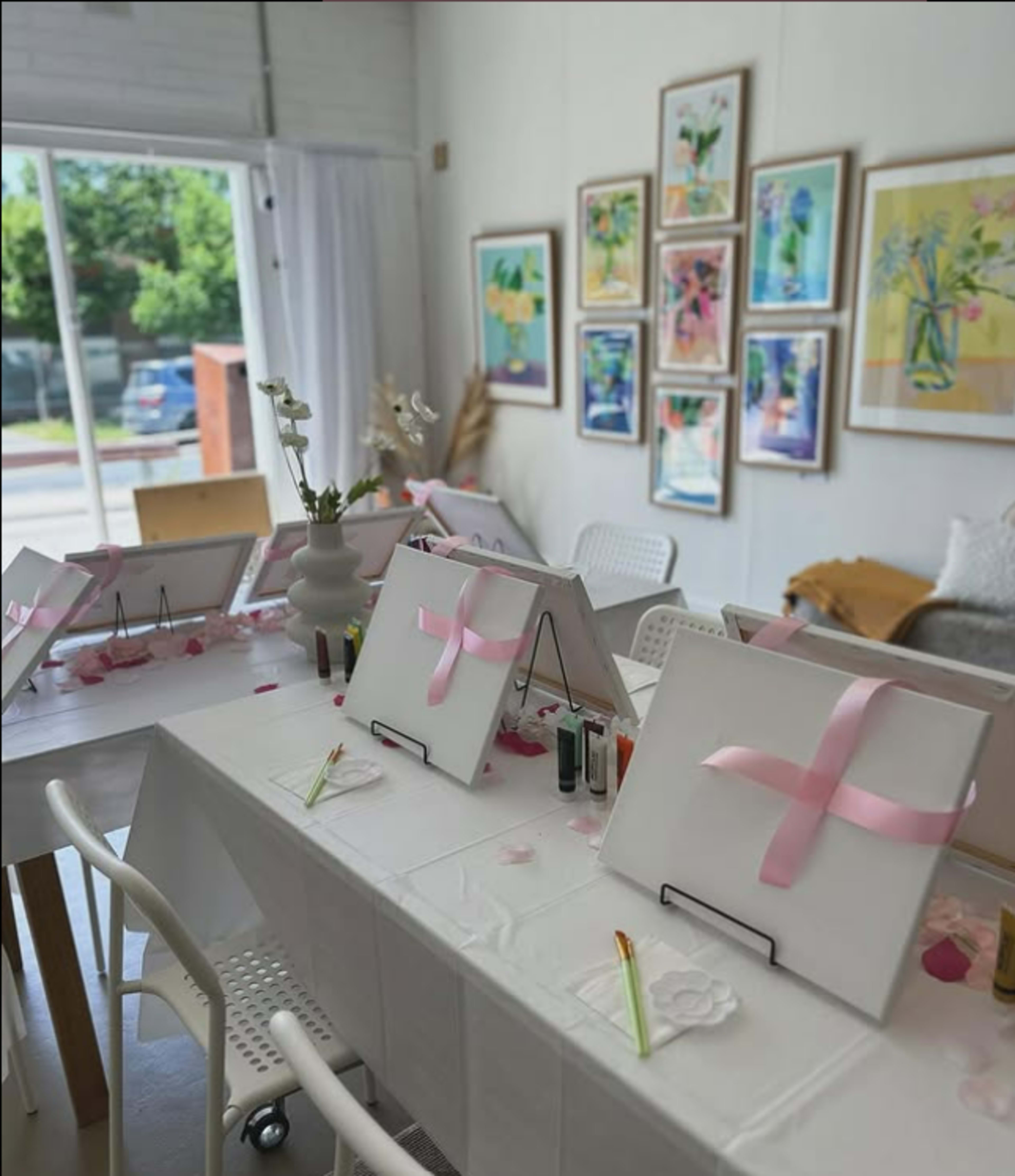 The image shows a bright indoor space set up for a painting activity, featuring several canvases adorned with pink ribbons on a long table surrounded by chairs.