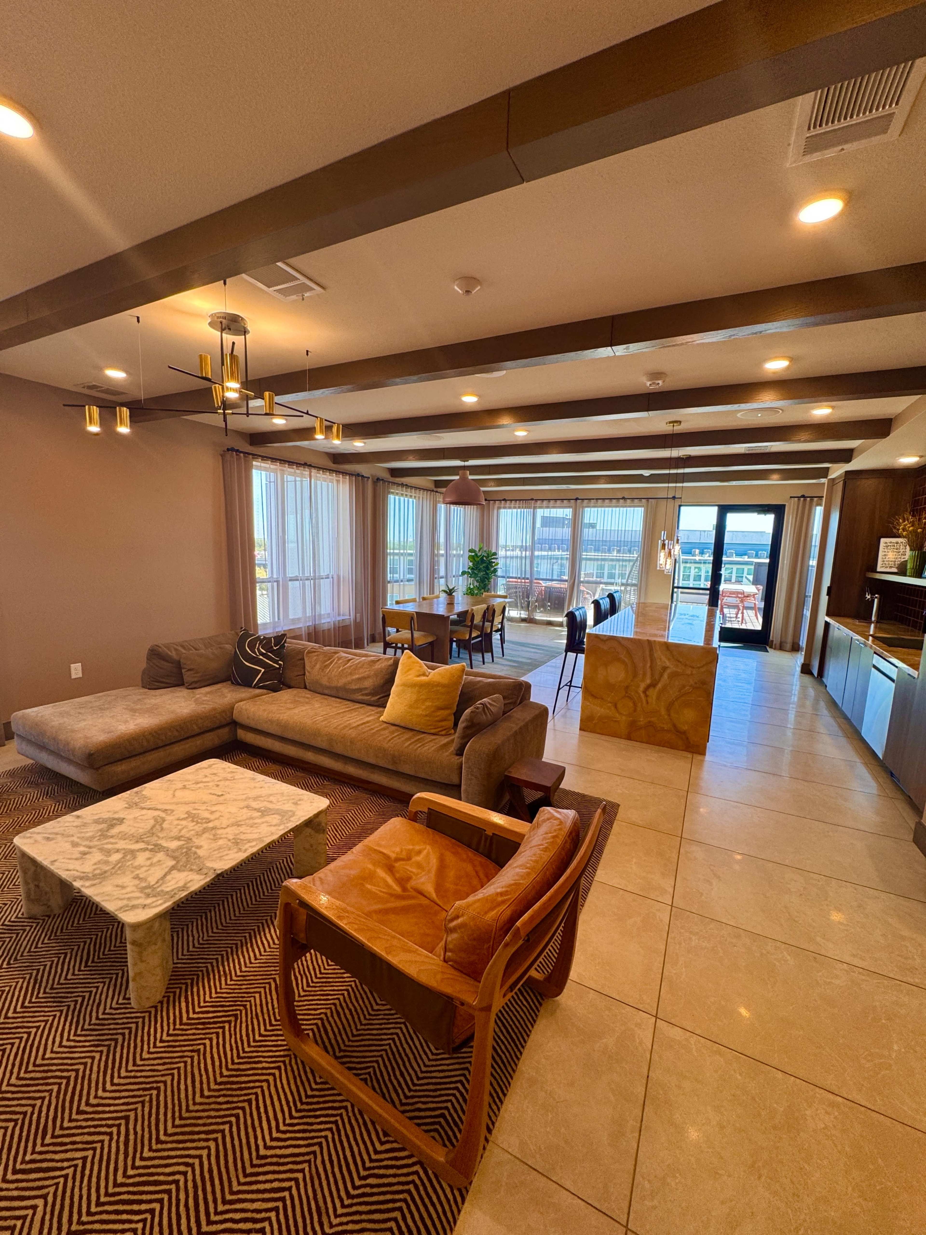 The image shows a modern living area with a sectional couch, a marble coffee table, and an armchair, set against a backdrop of large windows and wooden beam accents on the ceiling.