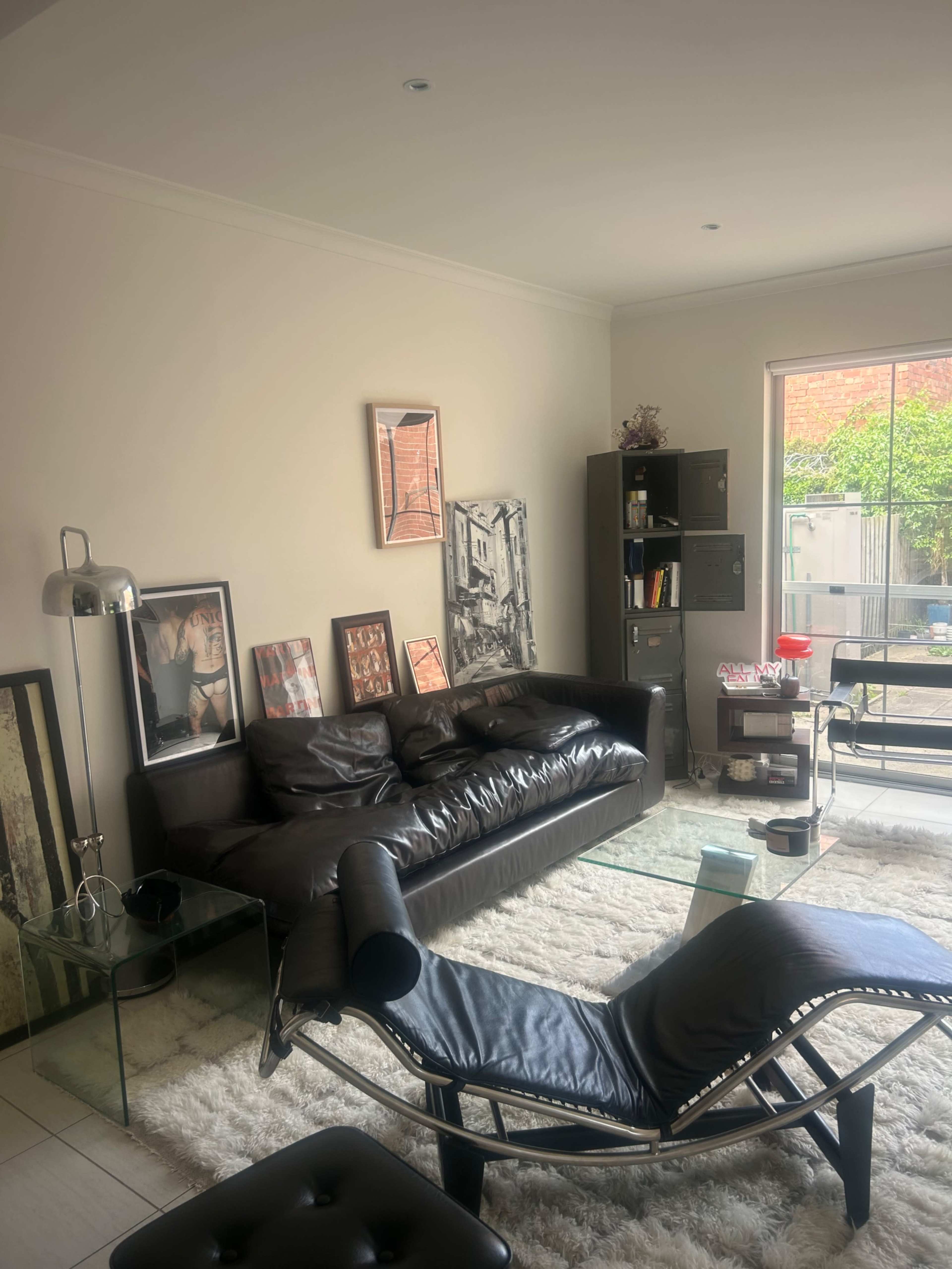 The image shows a modern living room with a black leather sofa, a glass coffee table, an armchair, several framed artworks on the wall, and a window revealing a patio area.