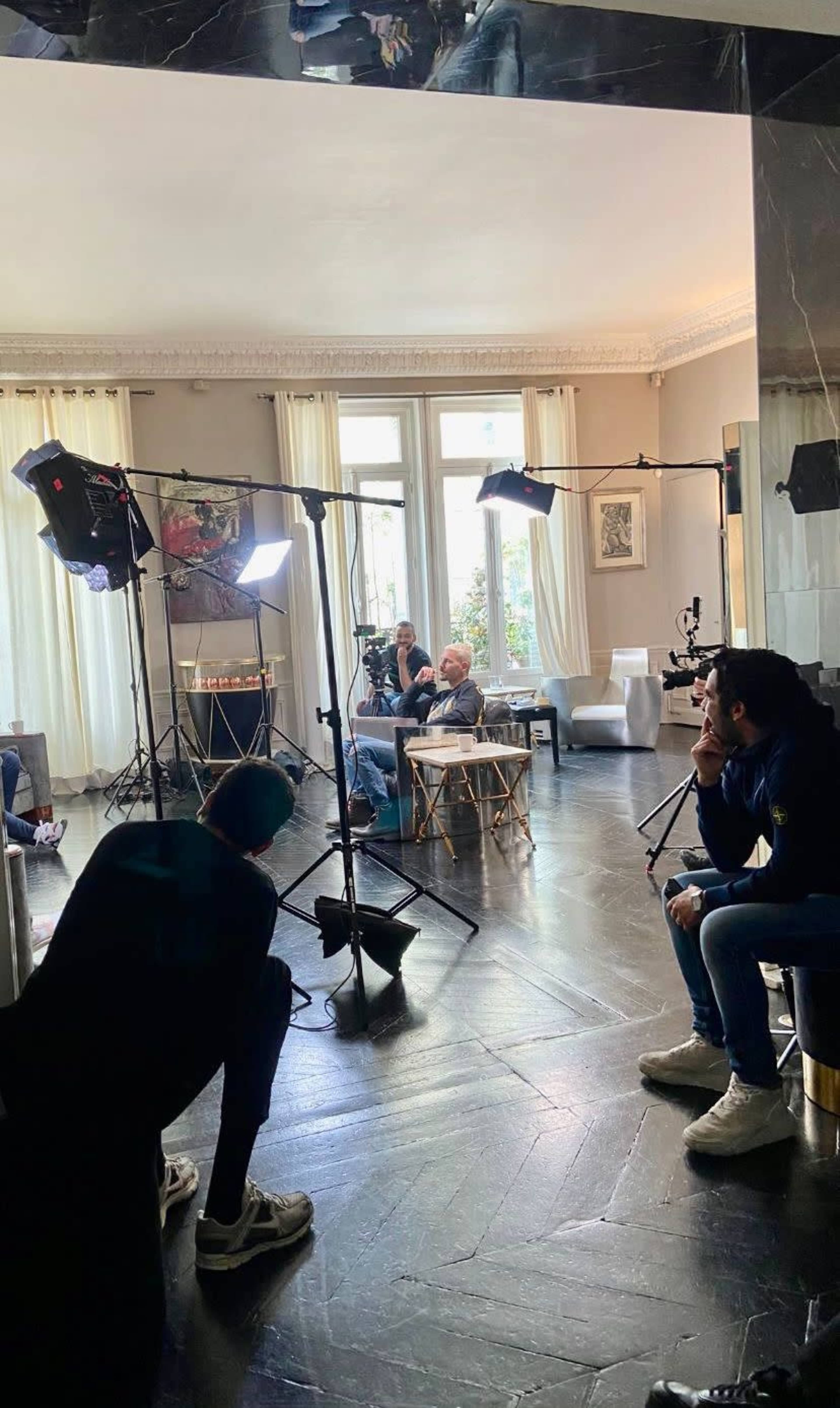 A group of people is sitting in a well-lit room with film equipment set up for a shoot.