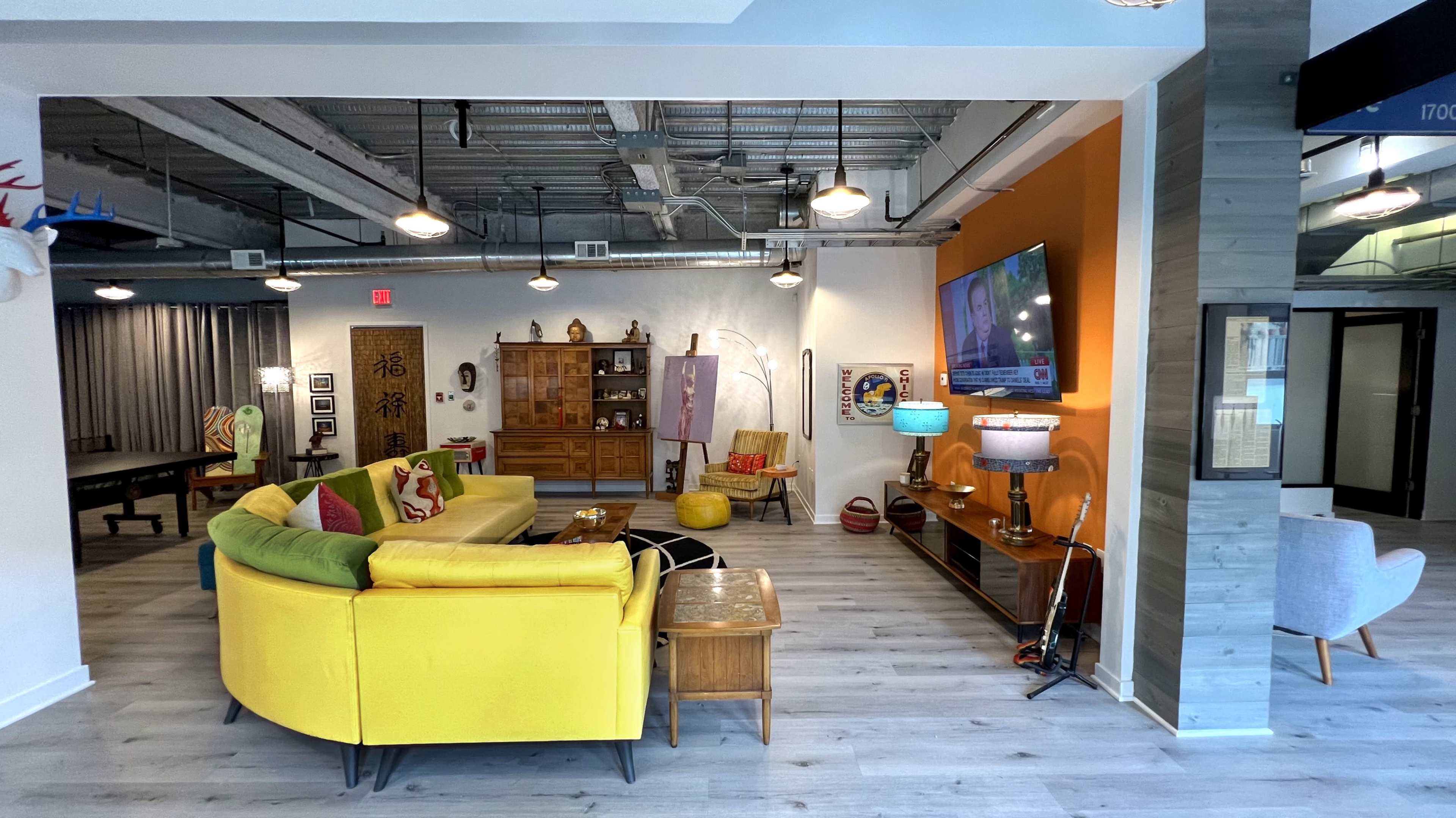 The image shows a modern, eclectic living space with a bright yellow sofa, a large television, and various decorative elements on the walls.