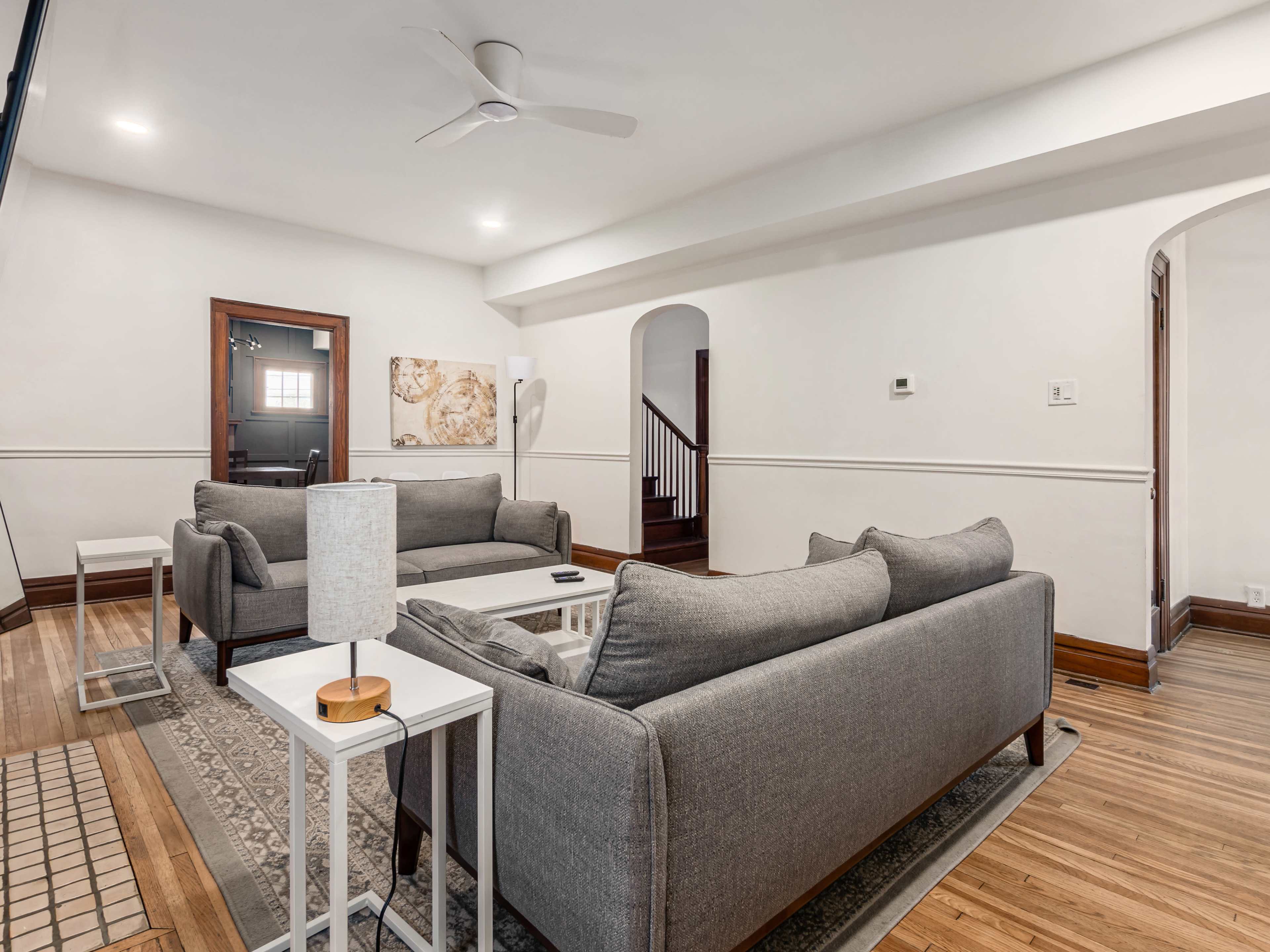A bright living room with two gray sofas, a white coffee table, and a ceiling fan, featuring wood accents and a staircase in the background.