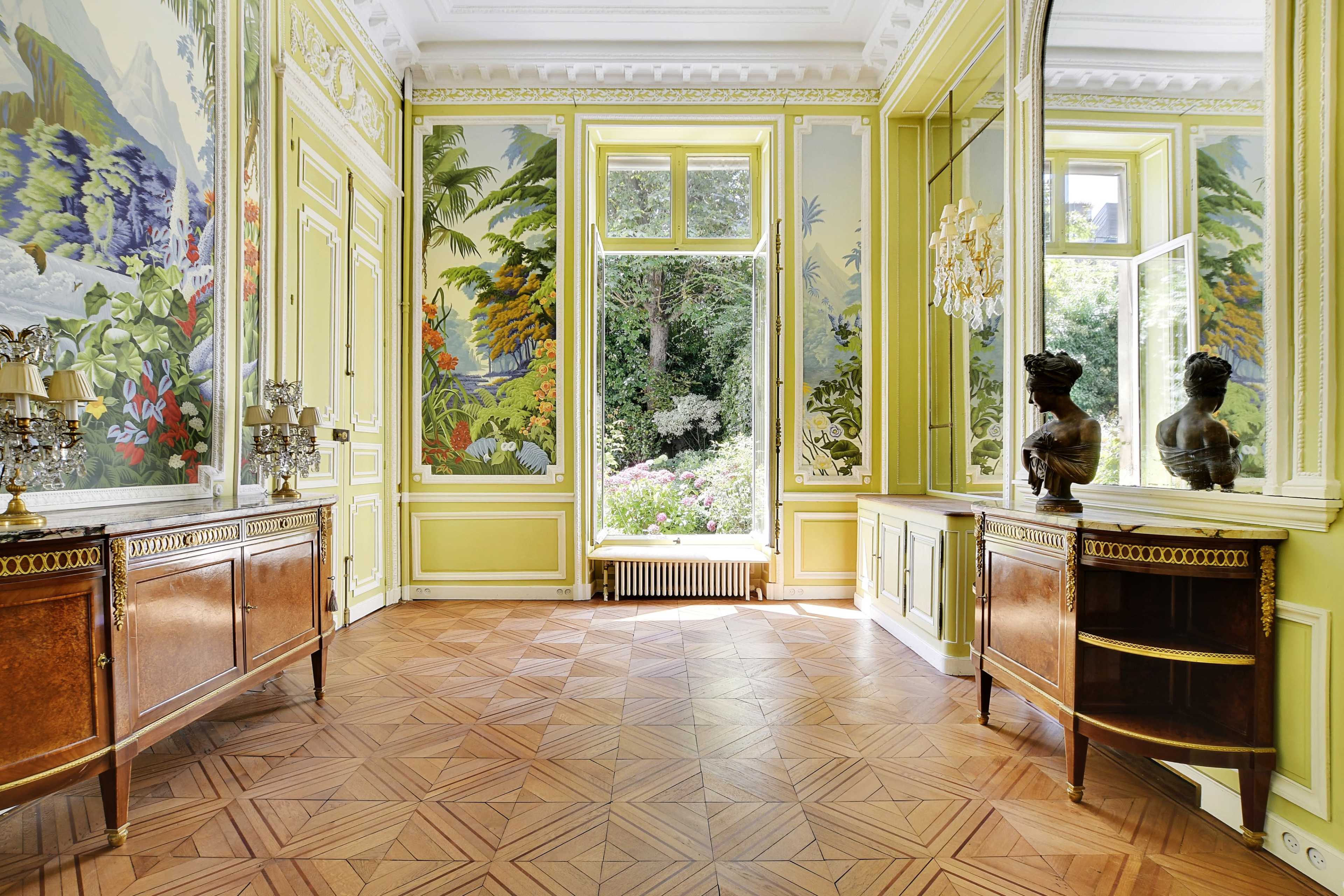 The room features elegant yellow walls adorned with botanical murals, large windows overlooking a garden, and wooden furniture arranged along the walls.