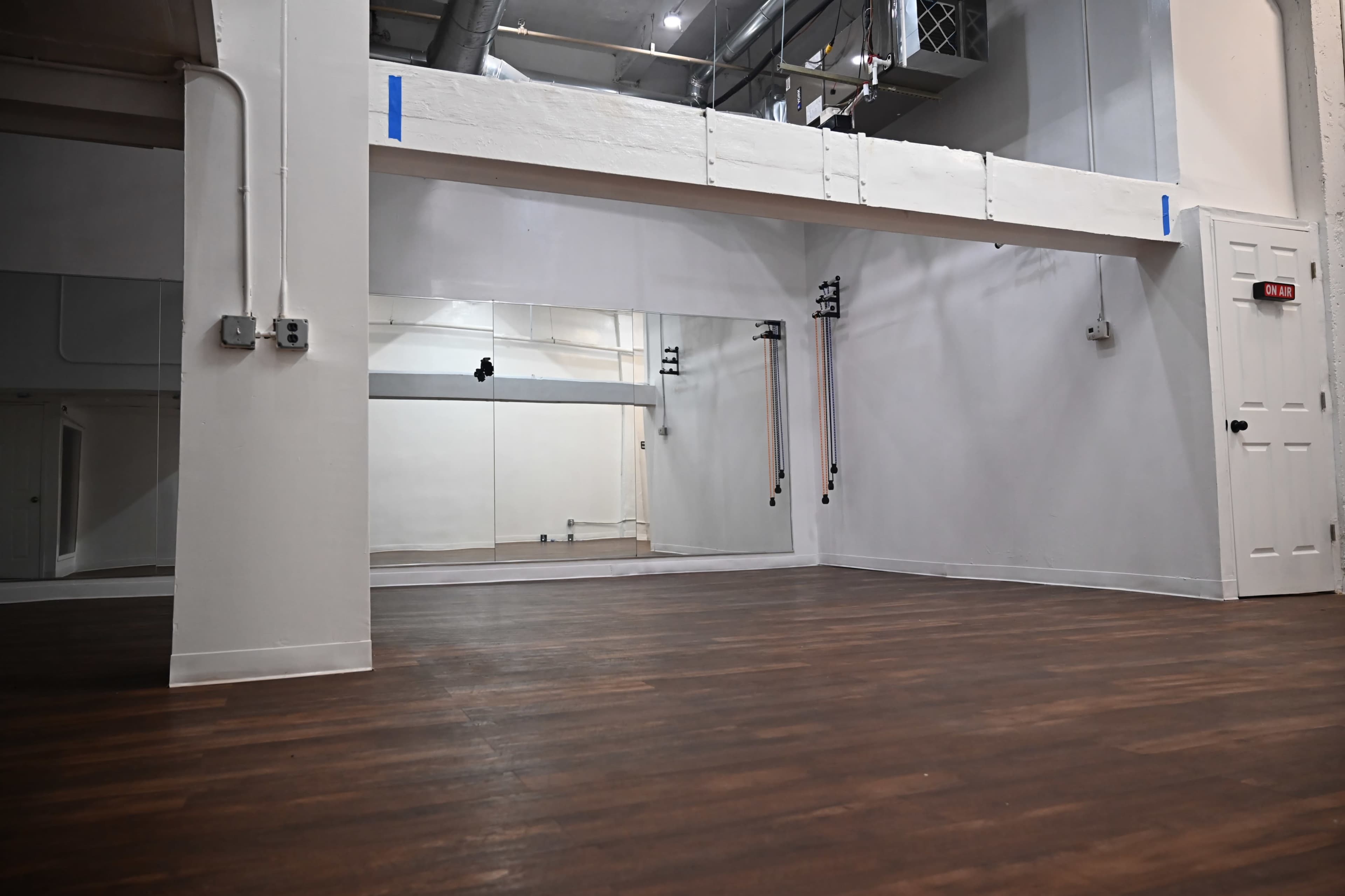 A spacious interior room with wooden flooring, white walls, and large mirrors, along with a door and exposed industrial elements in the ceiling.