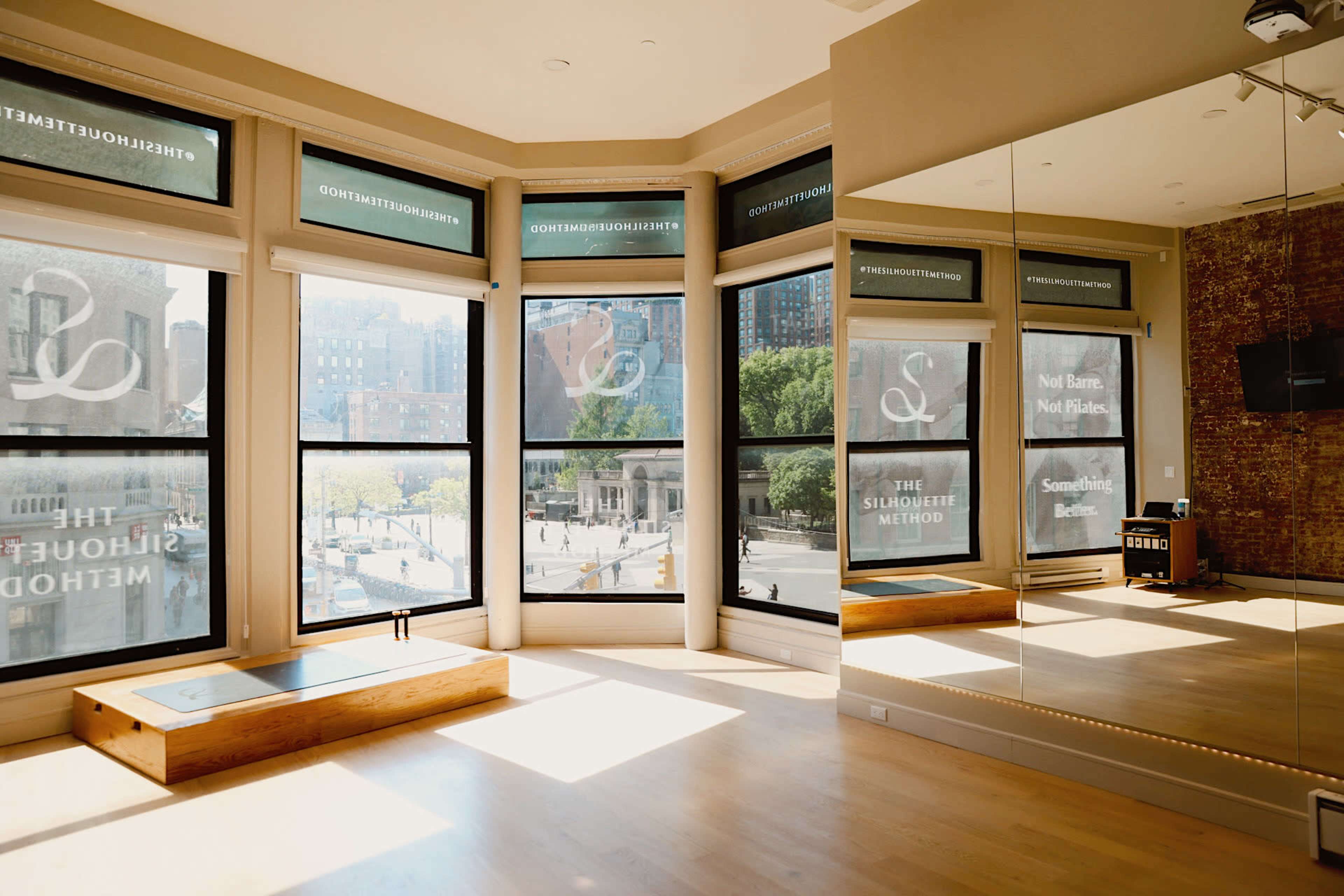The image shows a bright, spacious fitness studio with large windows overlooking a cityscape.